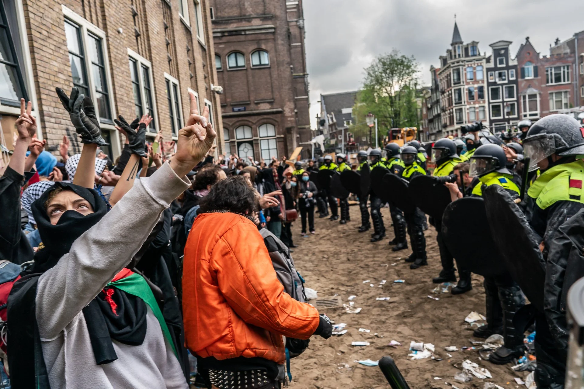 Protesters facing police officers in riot gear on a city street with brick buildings in the background. Some protesters have raised fists and make peace signs, others wear masks and hoodies. The ground has litter, and the atmosphere appears tense.