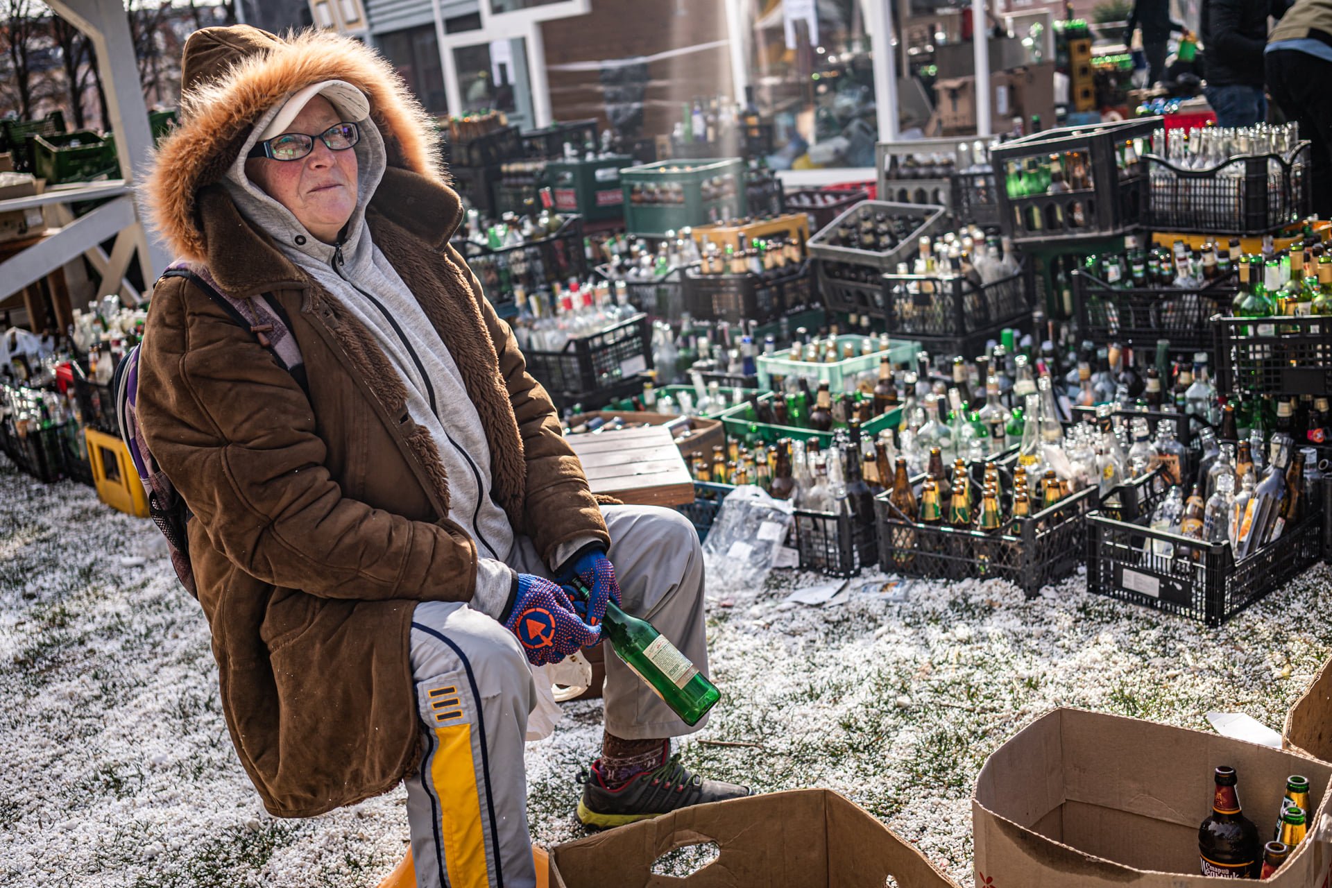 A person sitting outdoors in a winter jacket and headphones, surrounded by crates of empty glass bottles, with some bottles on the ground and in a cardboard box, during a cold time.