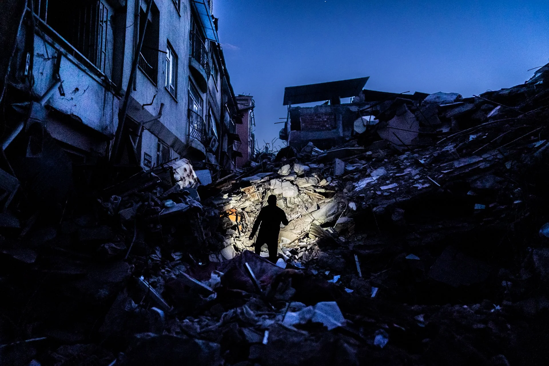 A person walking through a disaster site with collapsed buildings and debris at night, illuminated by a flashlight.