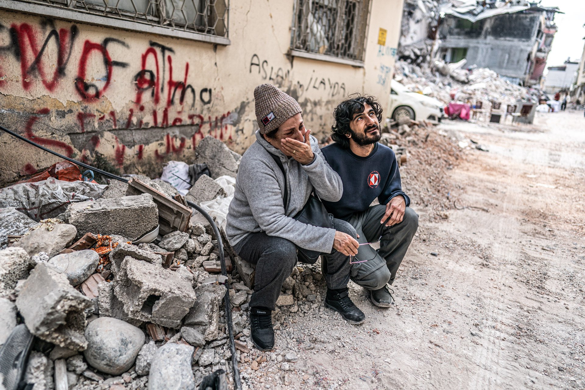 Two people sitting amidst rubble and debris in front of a damaged building, one appears distressed, covering their face; the other looks upward with a concerned expression.