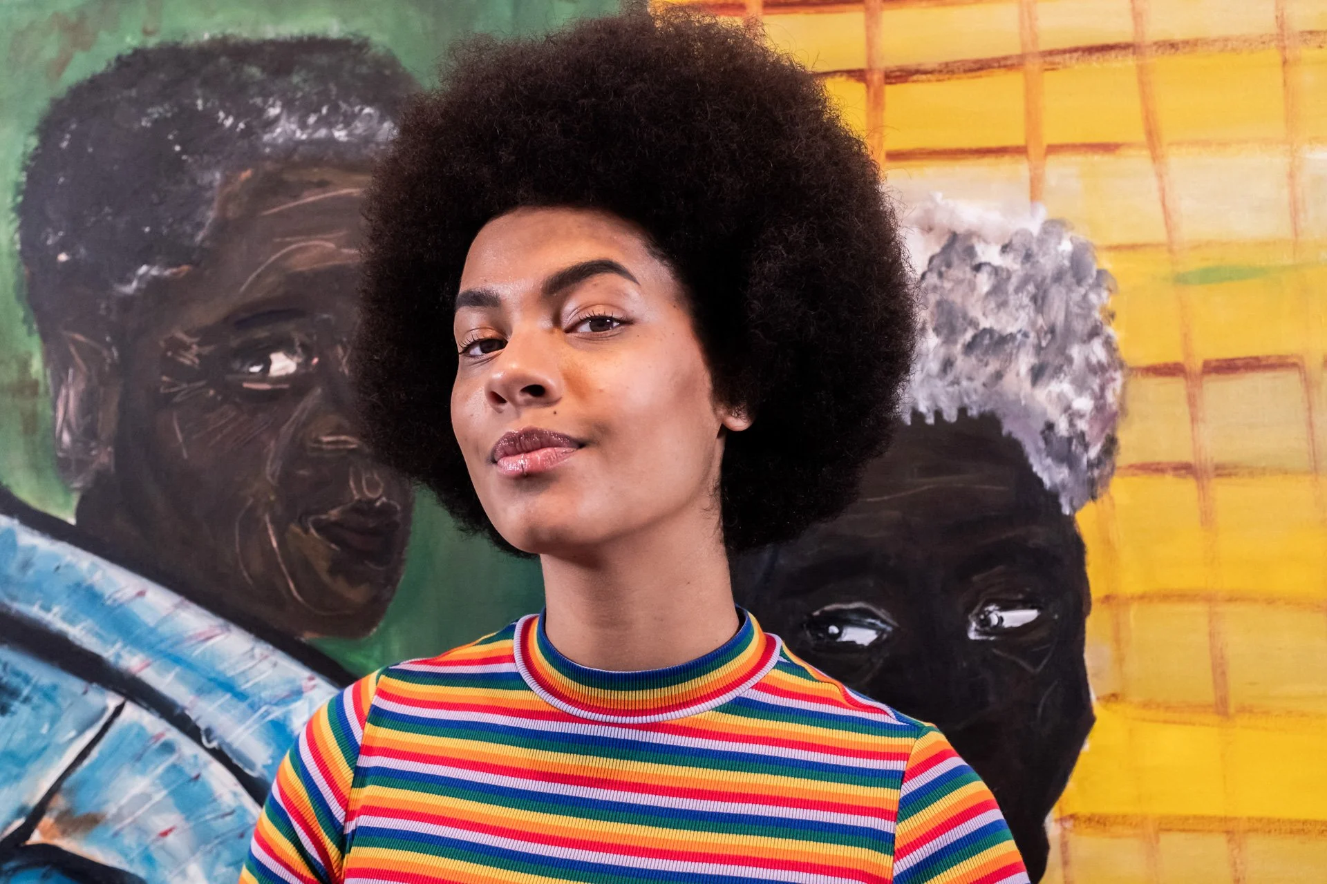 A woman with natural afro hair wearing a rainbow striped top, standing in front of a colorful mural featuring portraits of two women with dark skin.