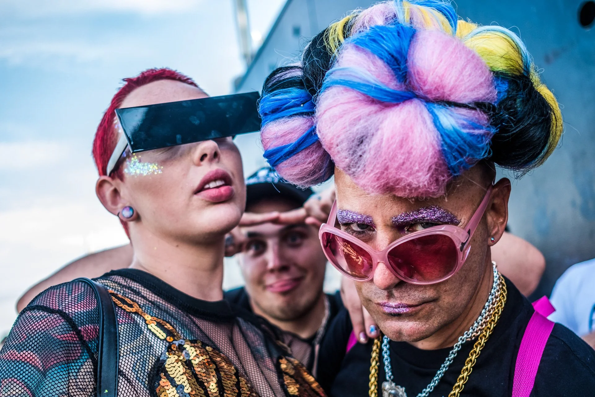 Three people dressed in vibrant, flamboyant clothing and accessories at a pride event. The person in the foreground has large, pink, heart-shaped sunglasses, glittery makeup, and colorful hair styled in large buns with pink, blue, yellow, and black c