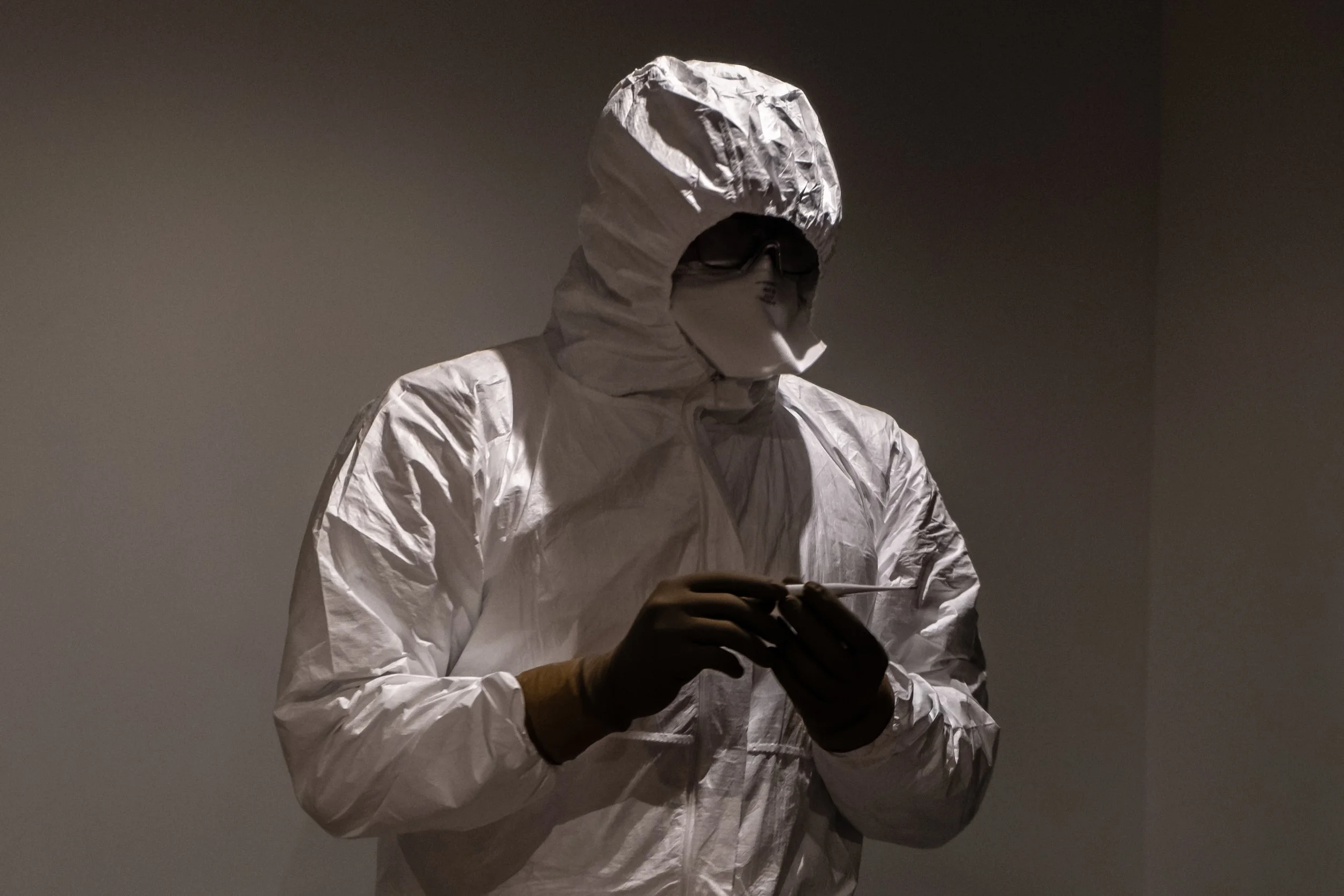 A person in a white hazmat suit and face mask holding a swab, standing in a dimly lit environment.