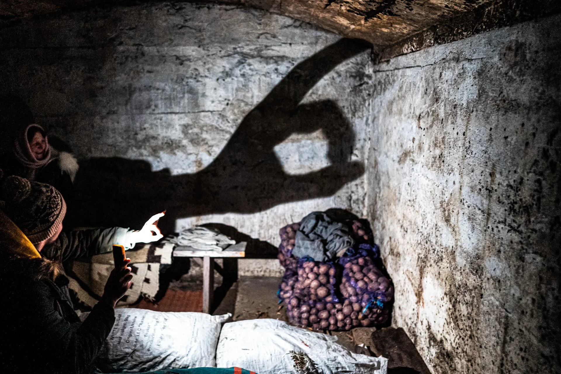 A group of people in winter clothing, with one person taking a photo of a shadow puppet on a rough stone wall using a mobile phone, with a pile of onions and some clothing on the ground nearby.