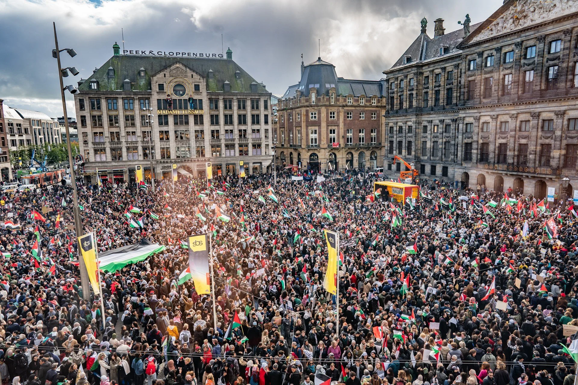 Large crowd of people gathered for a demonstration or protest in an urban square, with many holding flags and banners, surrounded by historic buildings.