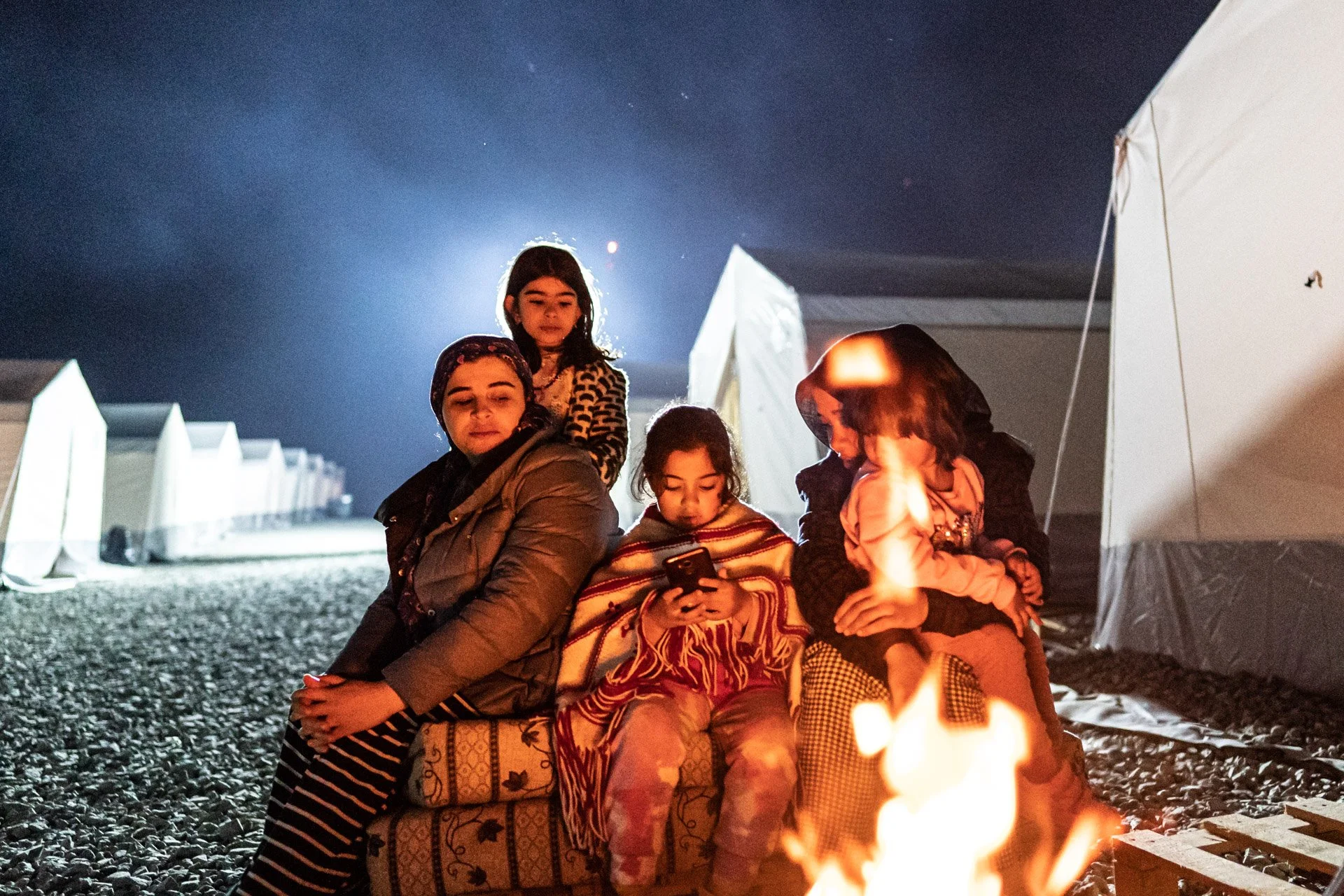Group of five children sitting around a campfire at night near tents, some looking at a phone, with a dark sky overhead.