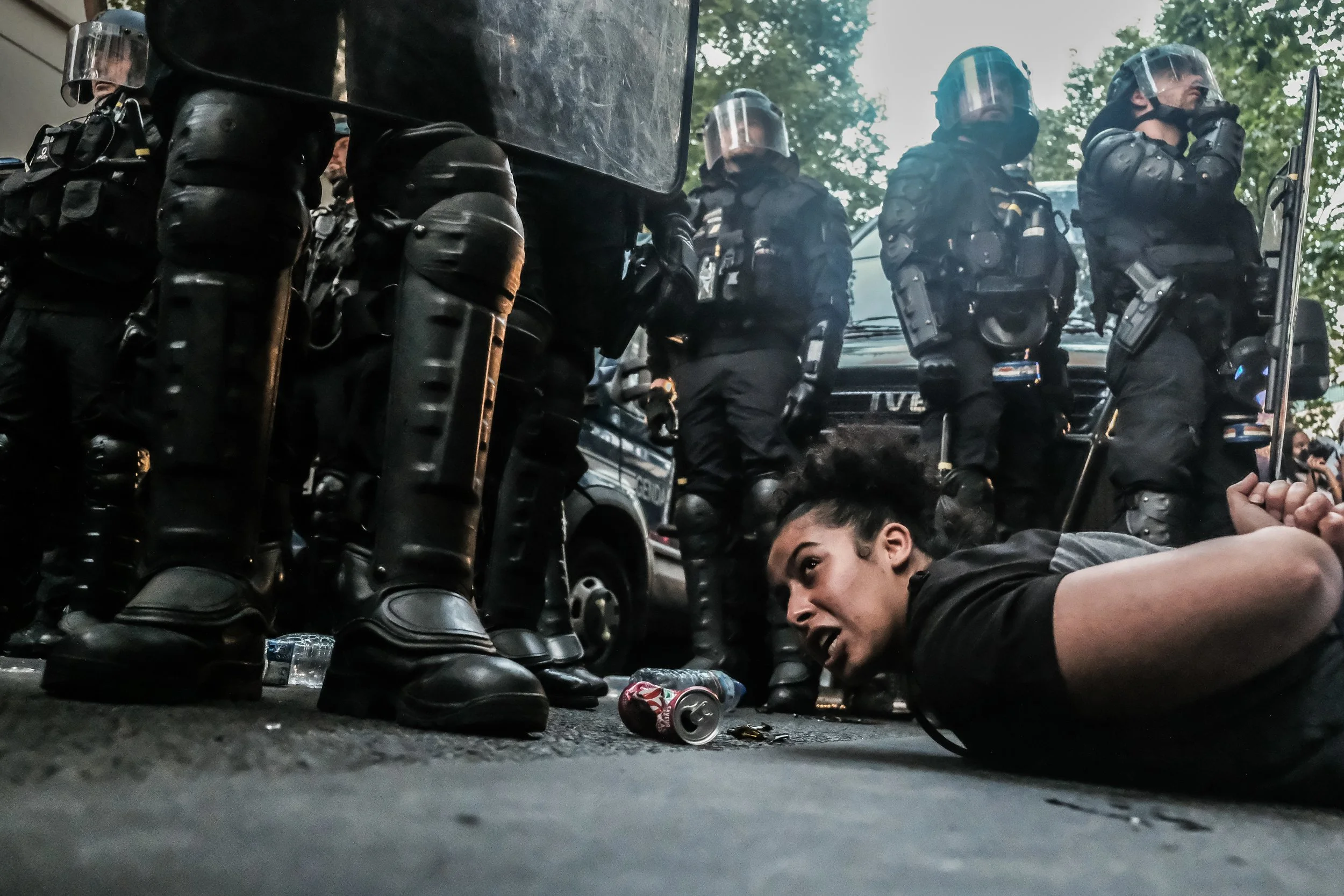 A woman is on the ground protesting with police officers in riot gear standing around her during a demonstration.