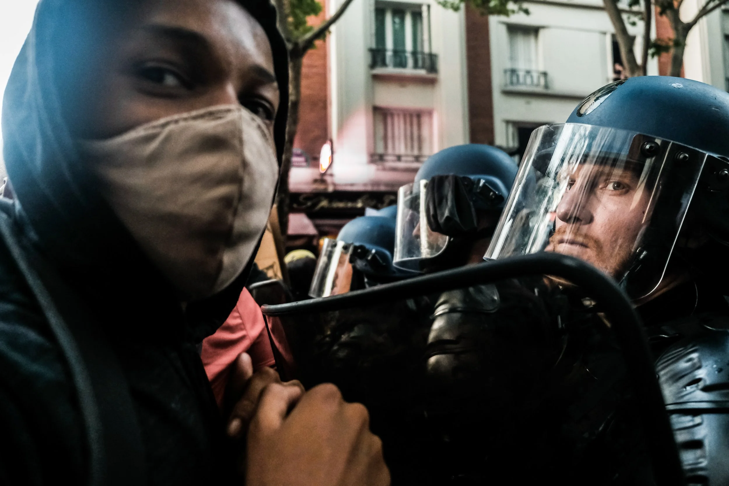 Close-up of a person wearing a beige face mask, standing next to police officers in riot gear with helmets and face shields, during daytime outdoors in an urban setting.