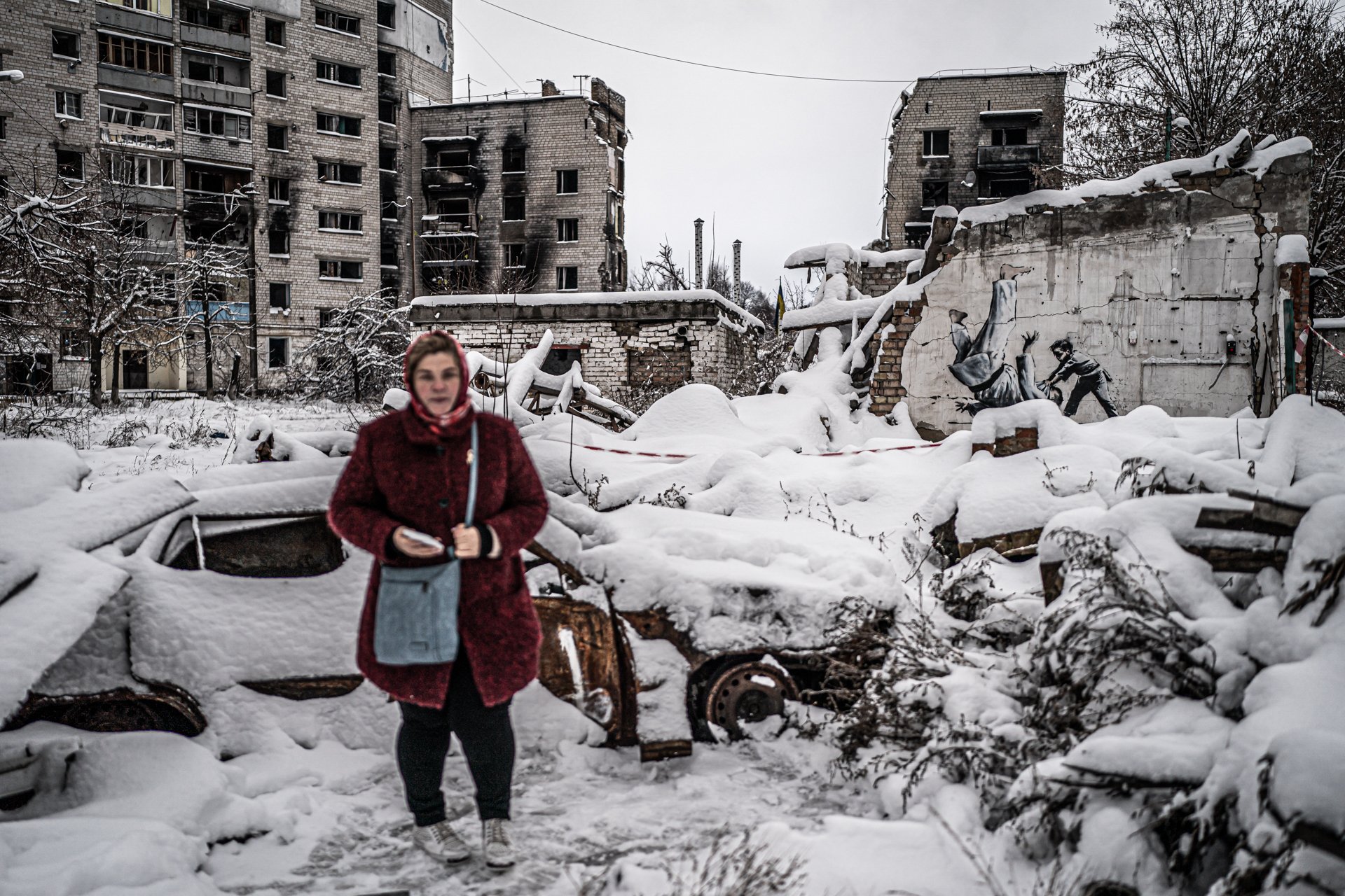 A woman stands in a snow-covered urban area with damaged buildings and debris. She wears a red coat and carries a gray bag, with a blurred face, amidst wreckage and abandoned cars, with a mural gunshot victim graffiti on a wall in the background.