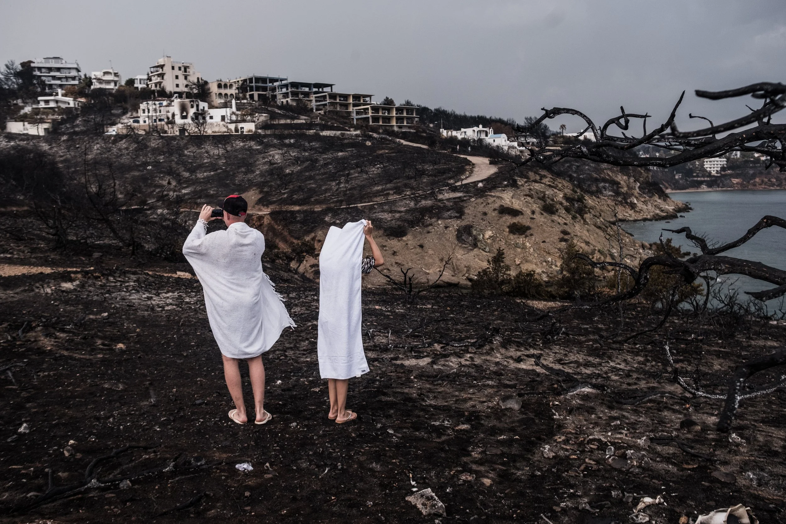 Two people dressed in white towels on a burnt landscape, one taking a photo and the other holding a towel, with a backdrop of burnt trees, hillside, and buildings under a cloudy sky.
