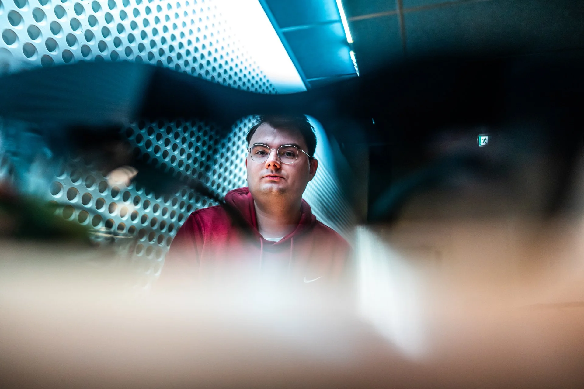 A man wearing glasses and a red hoodie sitting behind a perforated metal surface, viewed through a blurred foreground, with a background of a metallic wall with circular patterns and a small green emergency exit sign.