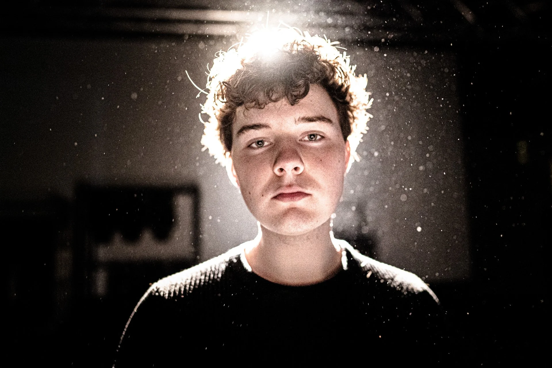 A young man with curly hair looking directly at the camera, backlit by a bright light creating a halo effect and dust particles in the air.