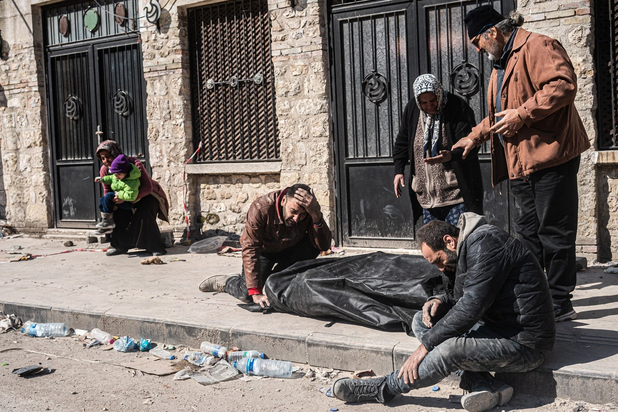 A group of people mourning on a street, with one person holding a child, and others sitting or standing around a covered body.