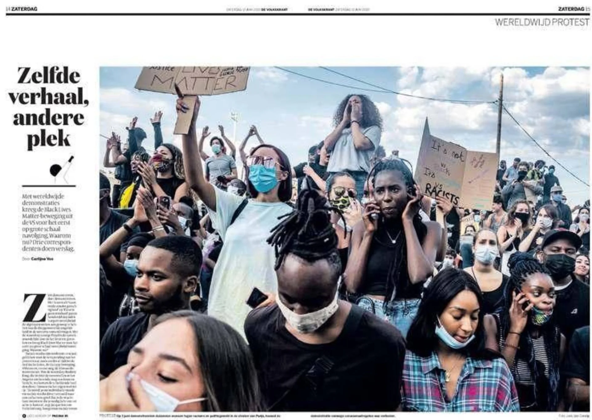 A diverse group of people protesting, many wearing face masks, some holding signs, with a woman raised hand and others on phones, against a partly cloudy sky.
