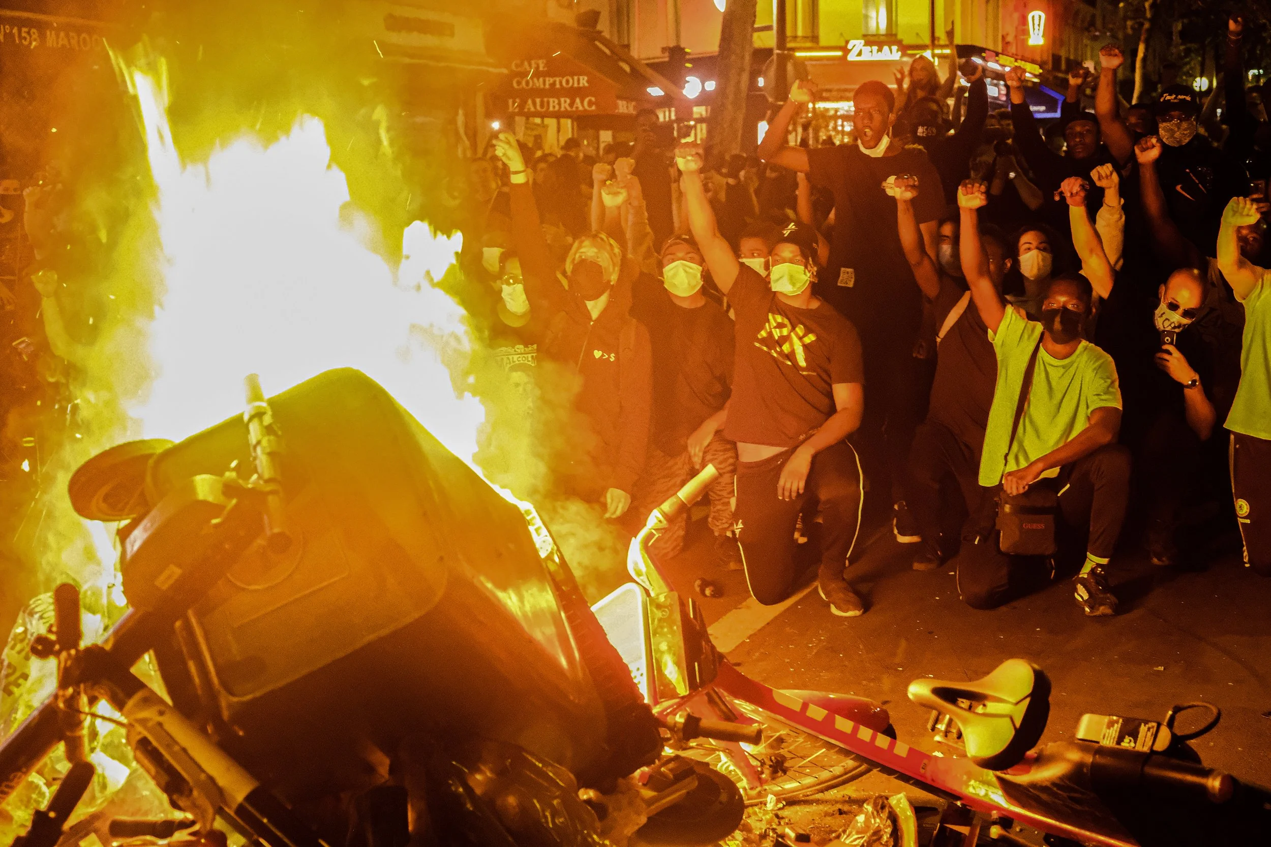 A group of protesters wearing masks kneeling and raising fists in front of a burning trash bin and overturned bicycle at night.