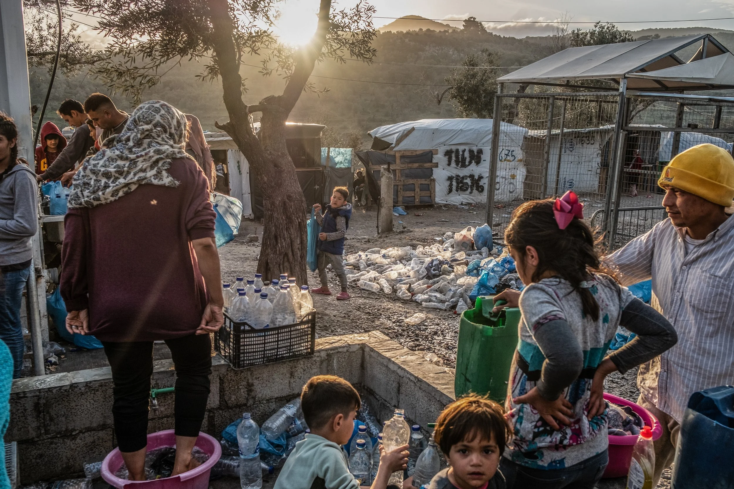 A group of refugees and children in a makeshift outdoor setting, collecting and sorting plastic bottles for recycling, with tents and a tree in the background, during late afternoon sunlight.
