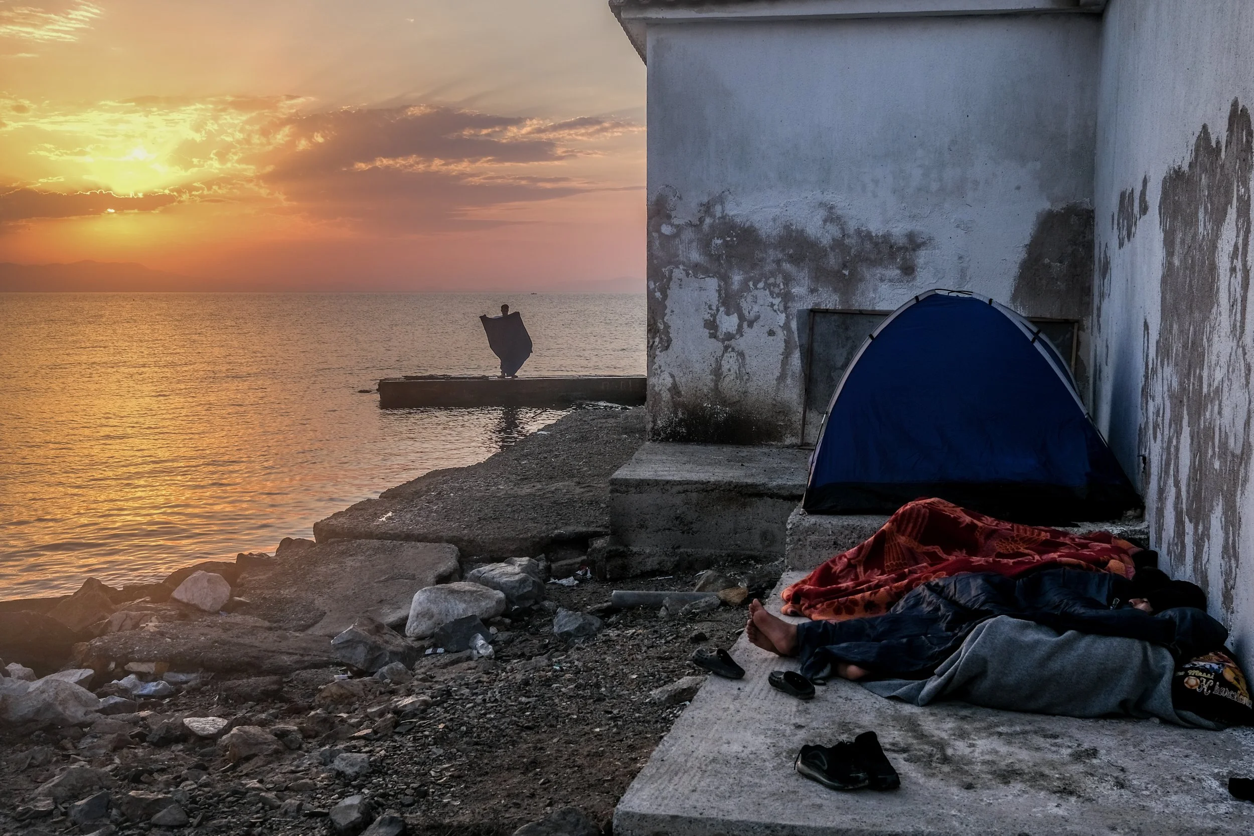 People sleeping on a concrete platform beside a weathered wall near the water at sunset, with a small tent and scattered shoes nearby.