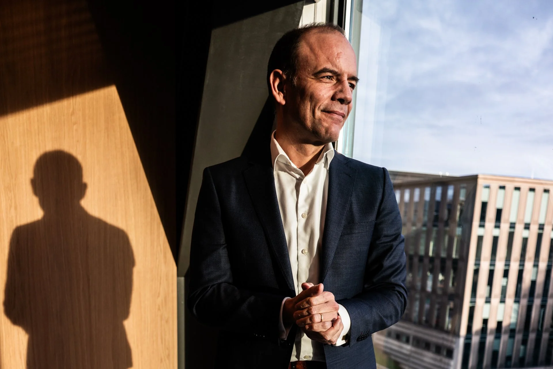 A man in a dark suit and white shirt stands near a window with sunlight shining on him, casting his shadow on the wooden wall behind him.