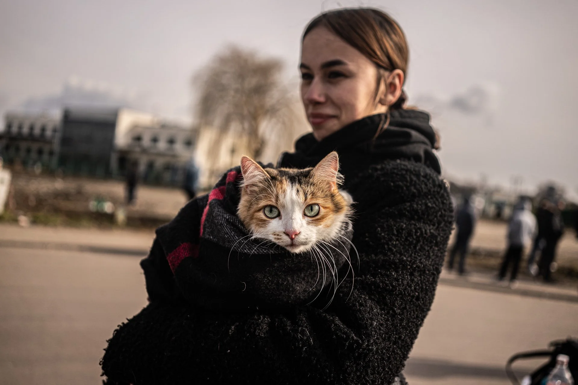A woman holding a tabby and white cat in her arms outdoors in a city park.