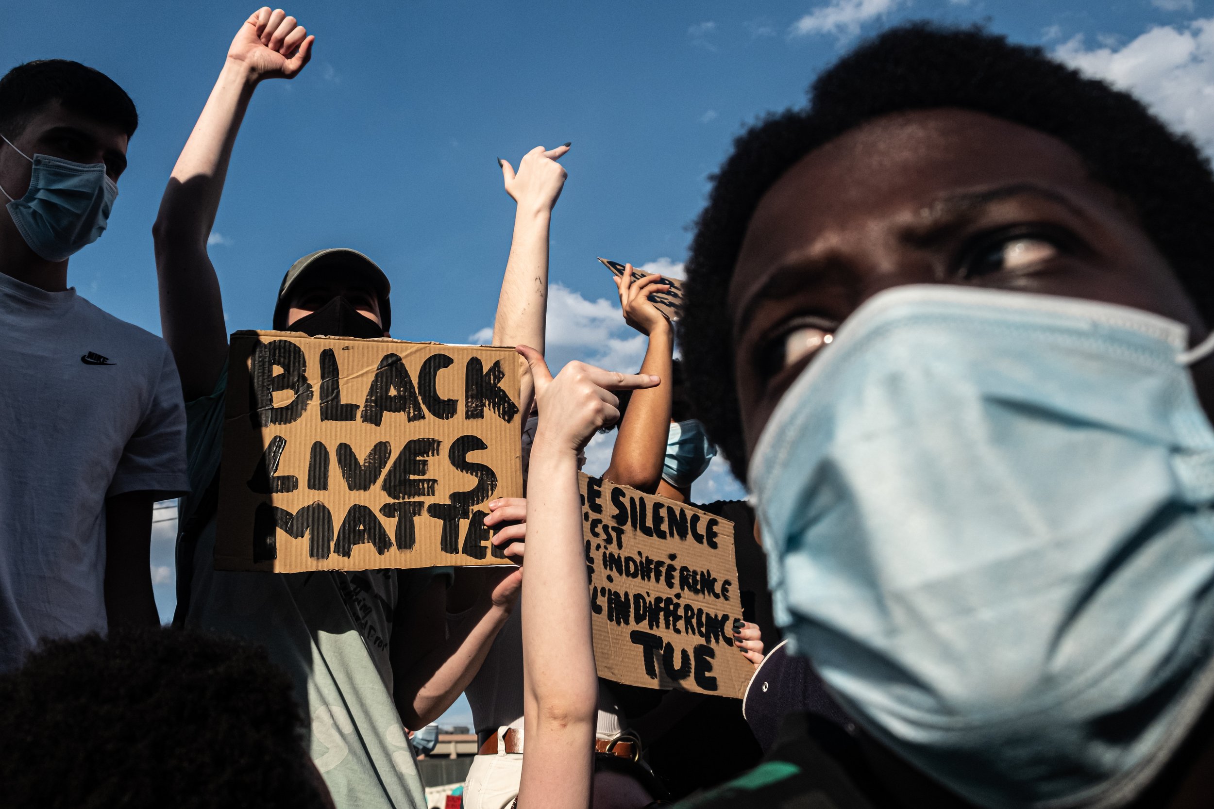 A diverse group of people wearing face masks participating in a protest, holding signs including one that says 'Black Lives Matter' and another with messages about silence and death.