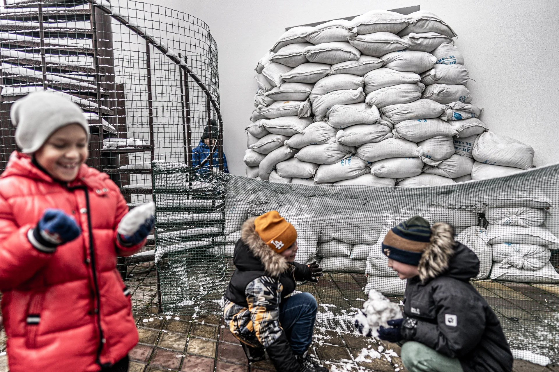 Children playing in the snow at an outdoor area with a wire fence, mound of sandbags stacked against a wall, and snow on the ground and nearby surfaces.