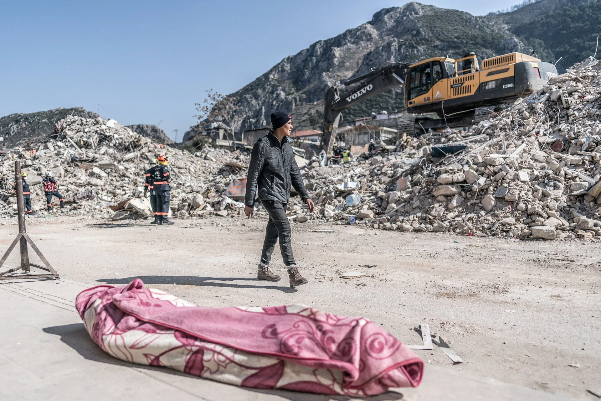 A person walks past debris and rubble from a collapsed building with a yellow excavator and rescue workers in the background. A pink folded blanket is on the ground in the foreground.
