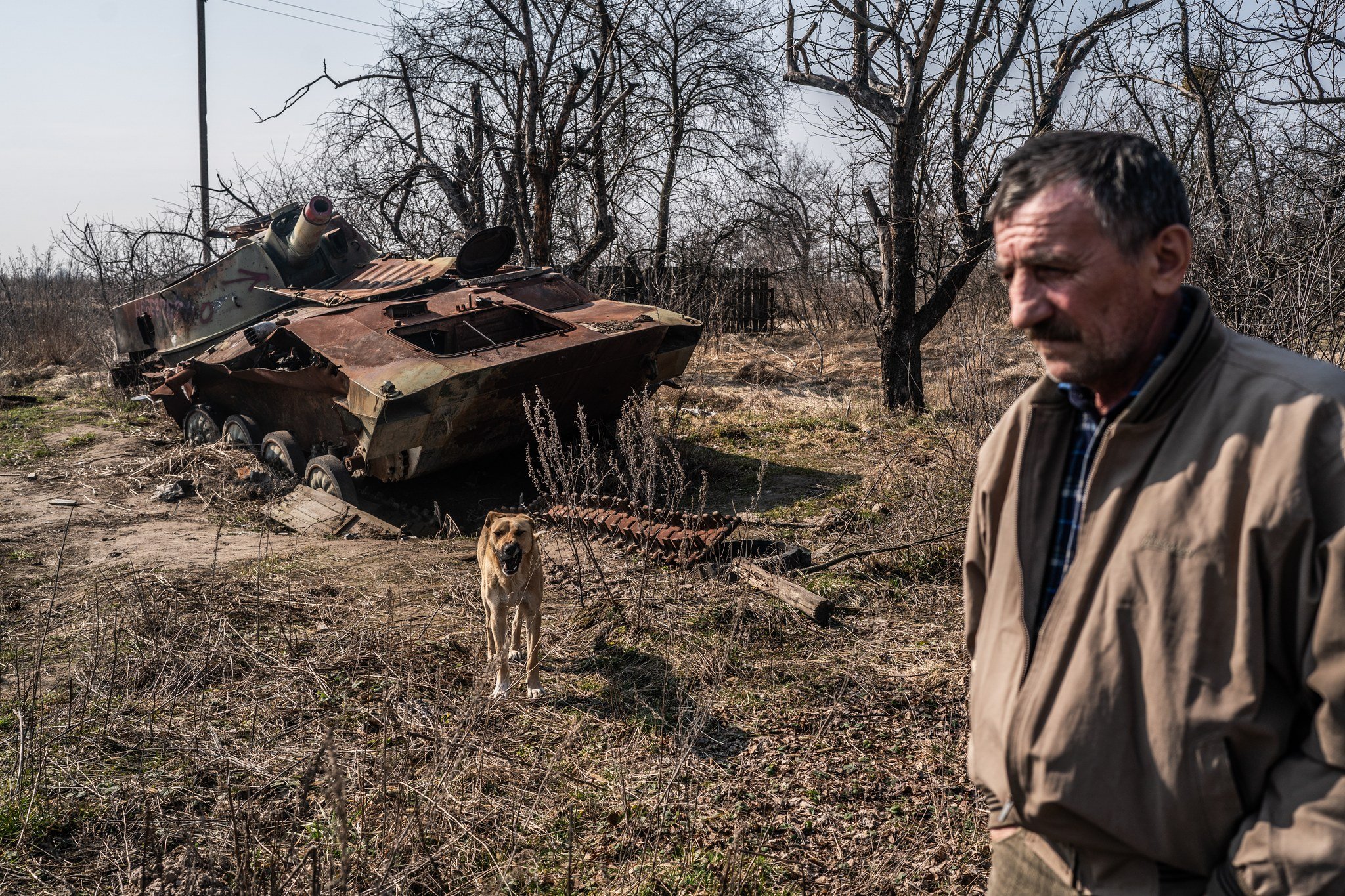A man standing in a field with a dog in front of an abandoned, rusty military tank with a destroyed turret, surrounded by leafless trees and dry grass.