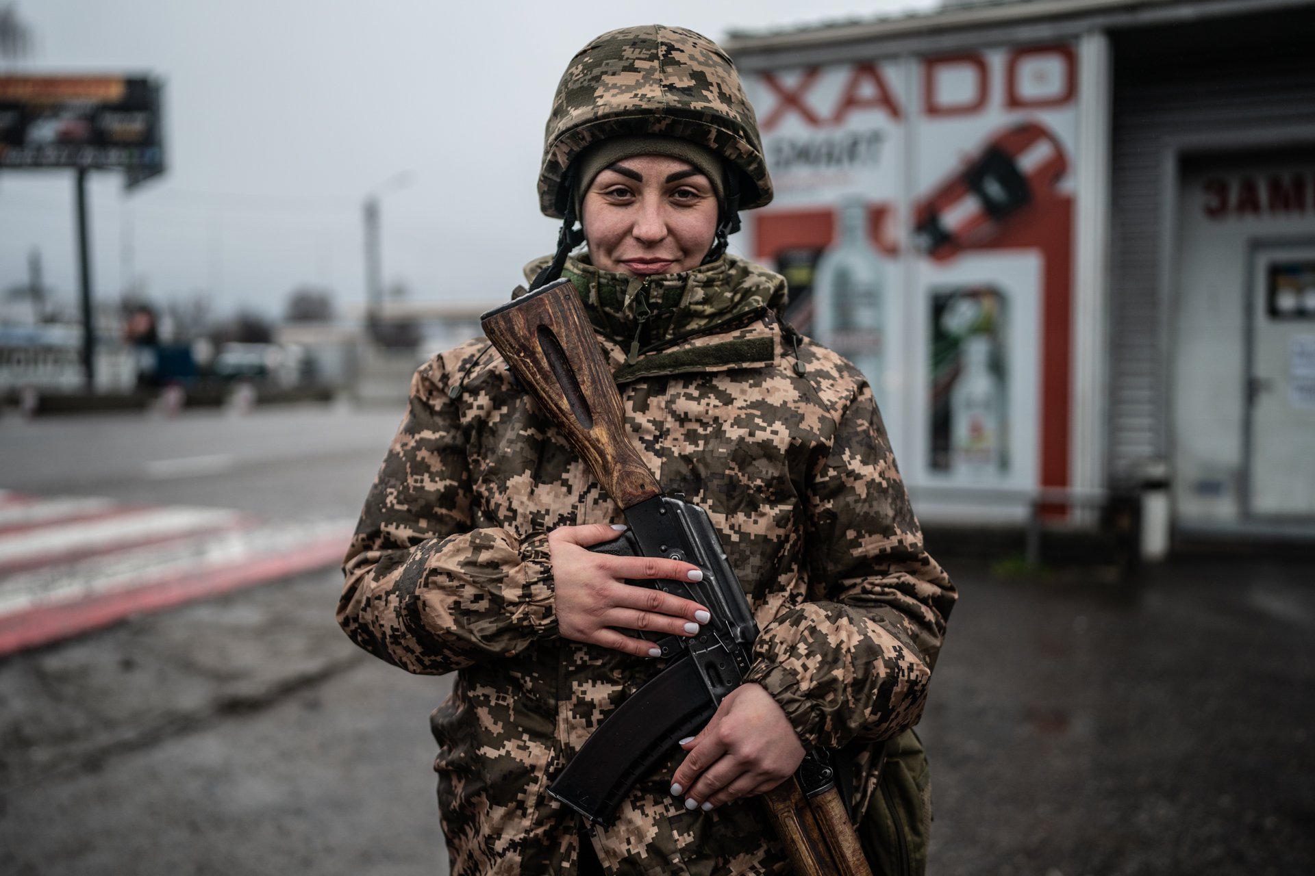 A female soldier in camouflage uniform holding a rifle, standing outdoors near a store with signs and advertisements in the background.