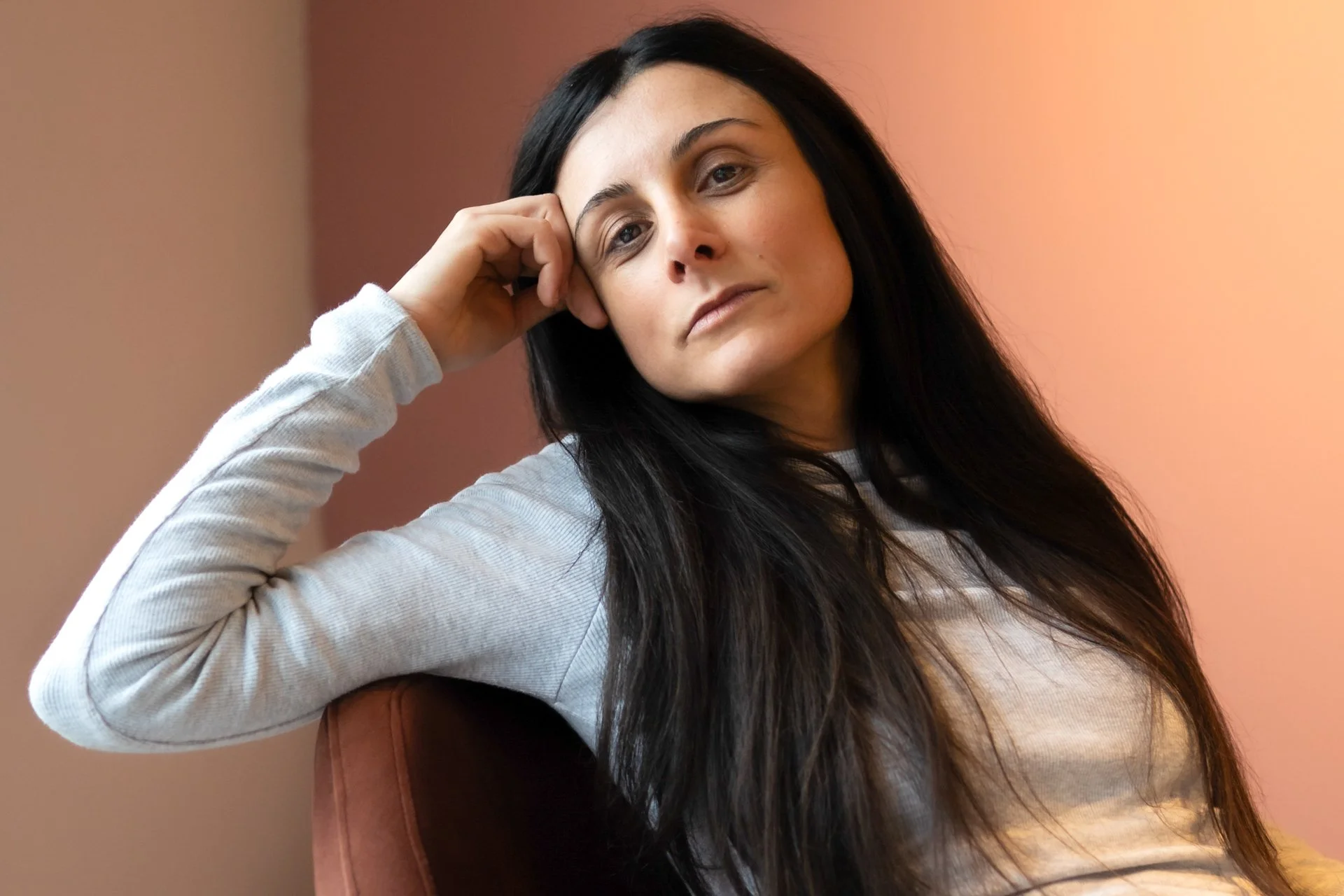 A woman with long dark hair resting her head on her hand, sitting against a pinkish background.