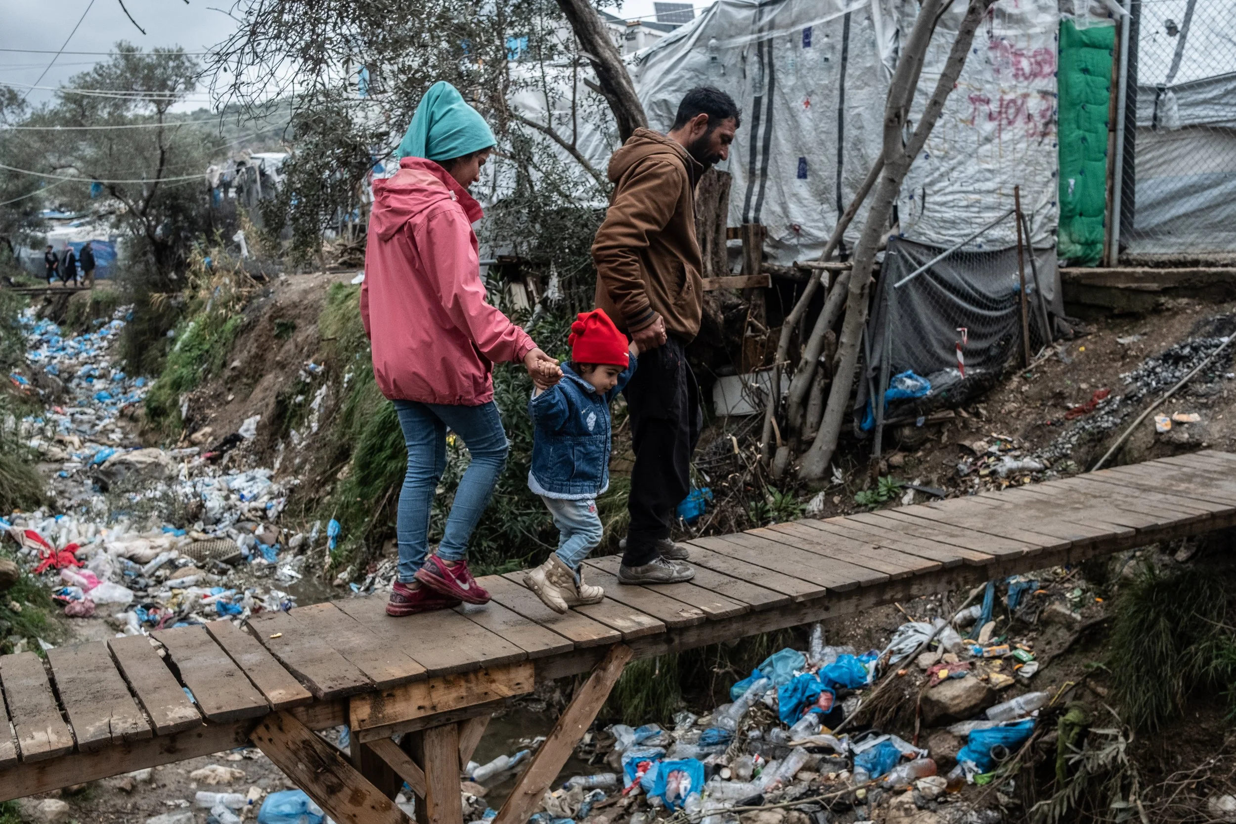 A family of three walking on a makeshift wooden bridge over a polluted river in a poor neighborhood. The family includes a woman, a man, and a young boy, all dressed warmly. The area around them is filled with trash and debris.