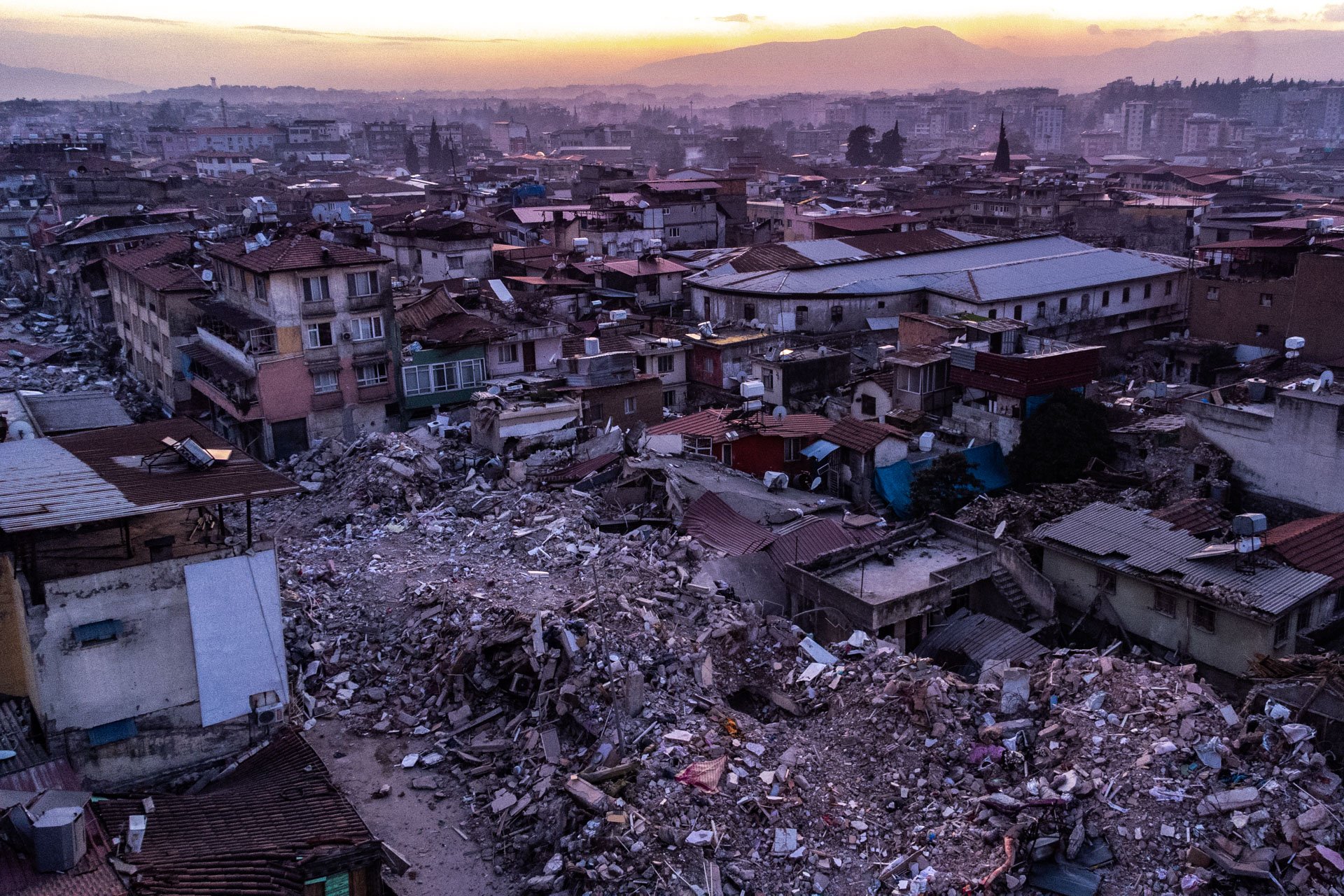 An aerial view of a city with numerous damaged buildings and debris, likely after an earthquake or disaster, at sunset with mountains in the background.