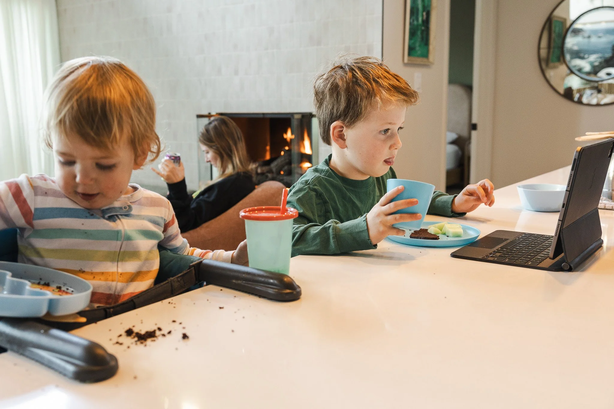 Two young brothers eat lunch at the kitchen counter while their mother relaxes by the fireplace in the background, an in-home documentary family photography session by Ruwan & Diana Photography, Del Mar.