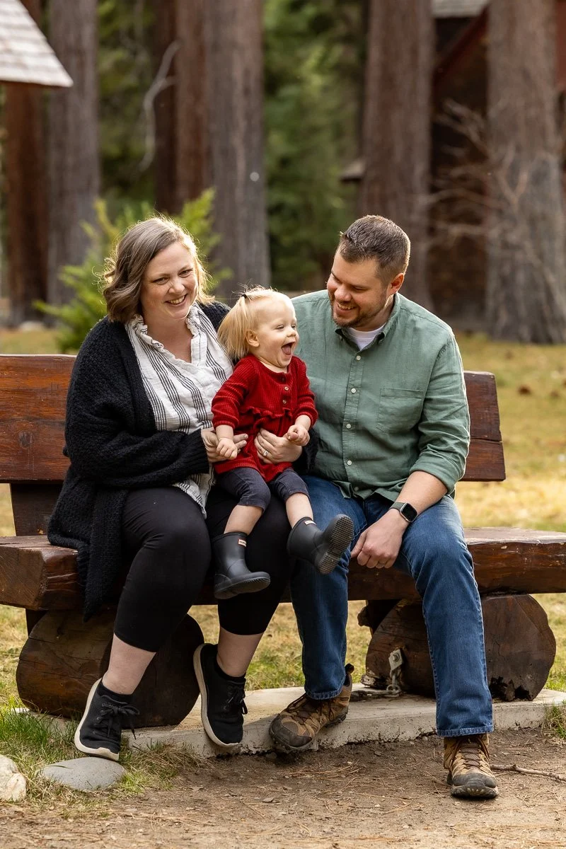Family sitting on a wooden bench at Camp Richardson with their toddler laughing between them during a documentary family photography session at Lake Tahoe