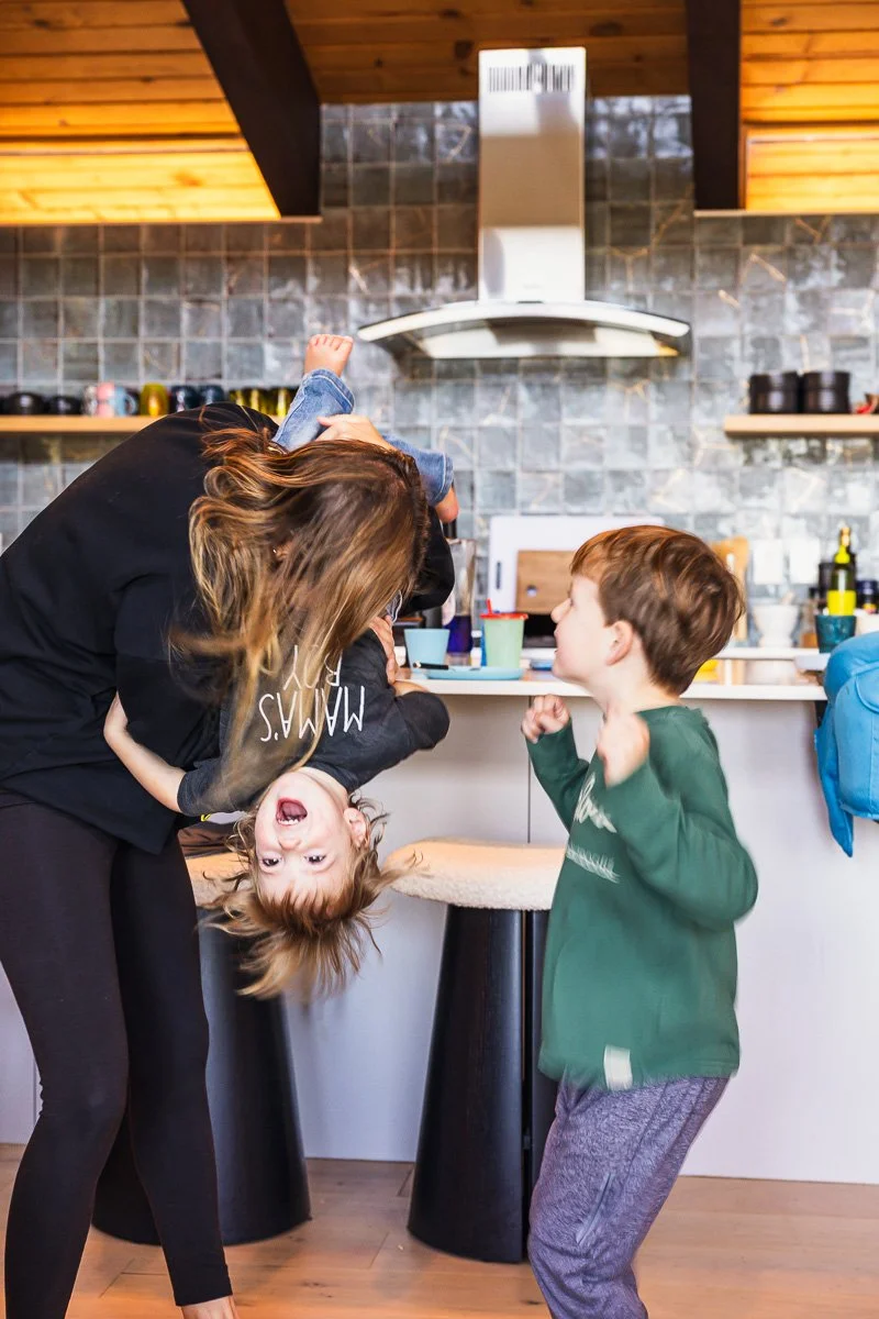  Mom swinging toddler upside down while older brother dances in cabin kitchen, Lake Tahoe, documentary family photography 