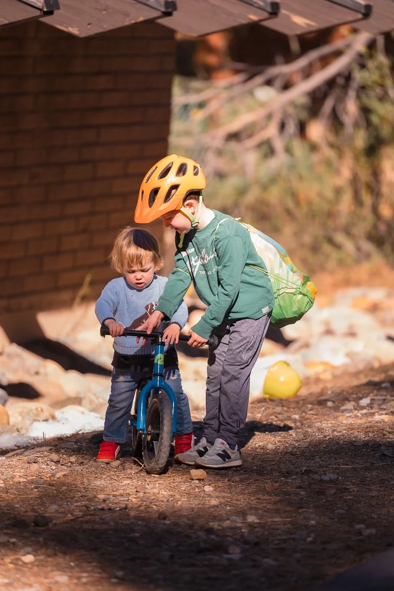  Older brother helping toddler onto balance bike on gravel path, Lake Tahoe, documentary family photography 