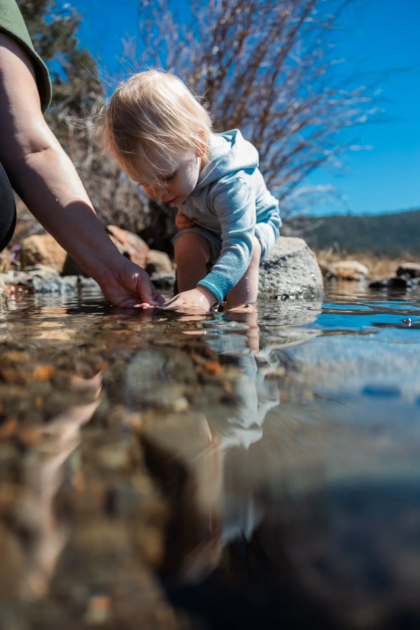 Toddler reaching for rocks in Fallen Leaf Lake shallows with mother's hand beside her, documentary family photography