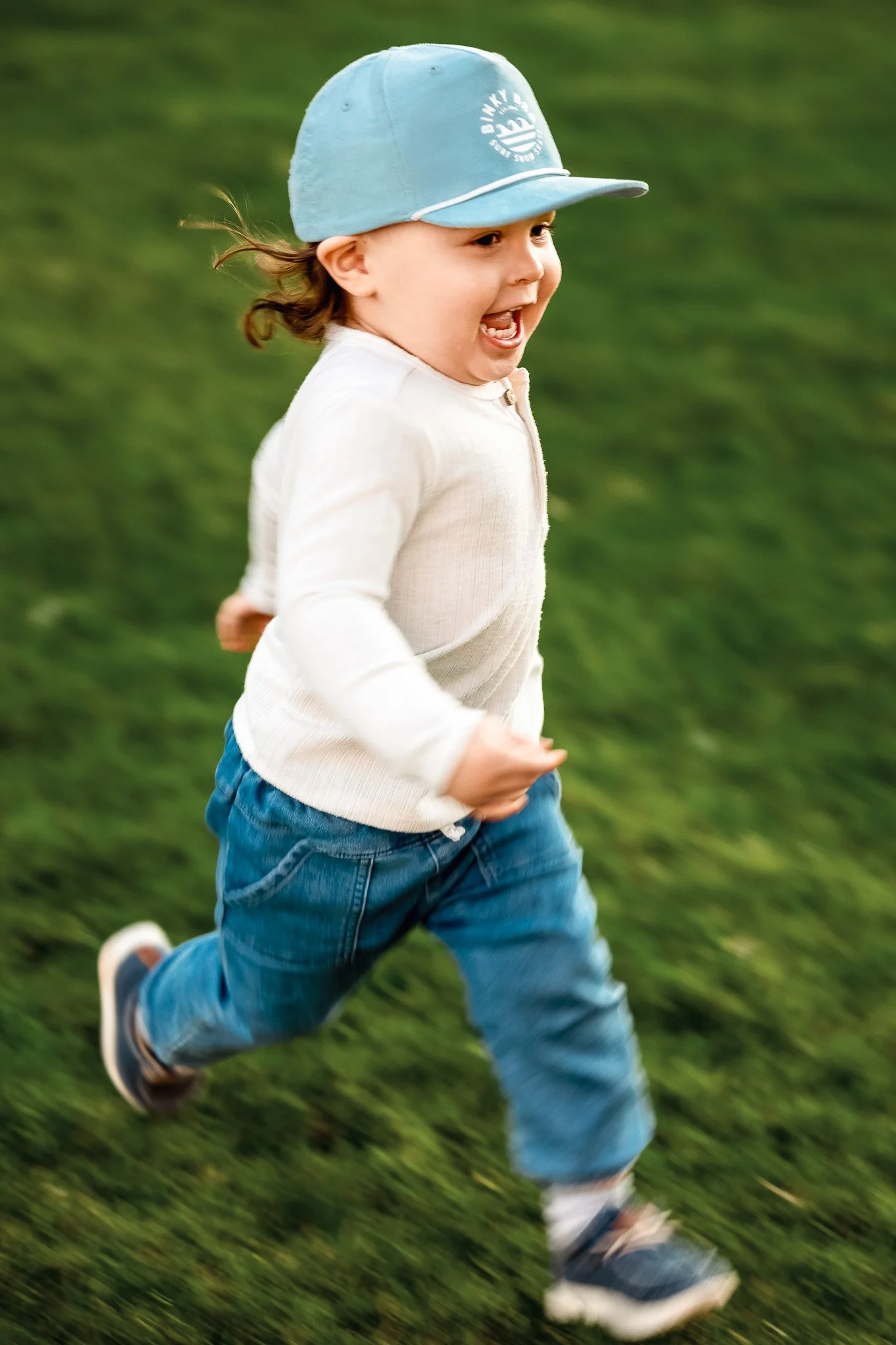 Toddler running through grass laughing in blue cap and jeans, documentary family photography San Diego