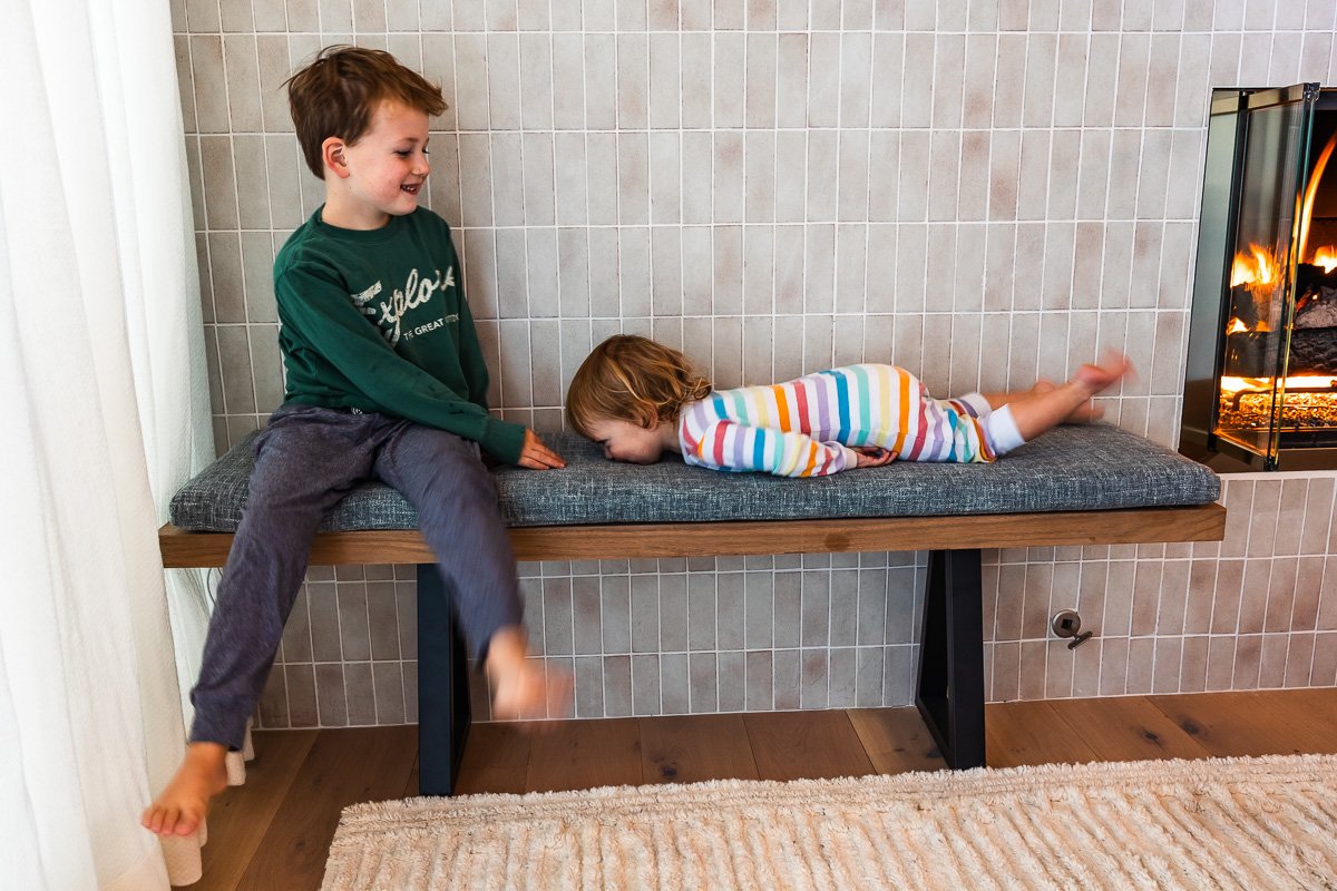 Older brother laughing as toddler face-plants on bench by fireplace, Lake Tahoe cabin, documentary family photography