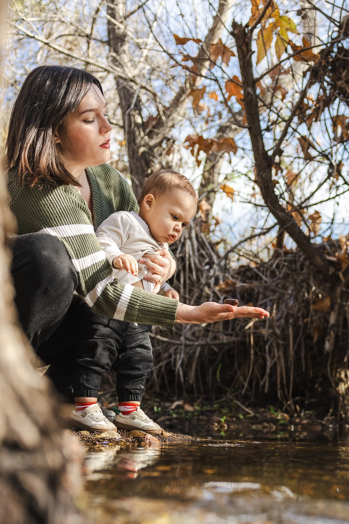 A mother crouches at the edge of a creek, holding a small stone in her palm for her baby to examine, a candid moment from a documentary family photography session by Ruwan & Diana Photography, Del Mar.