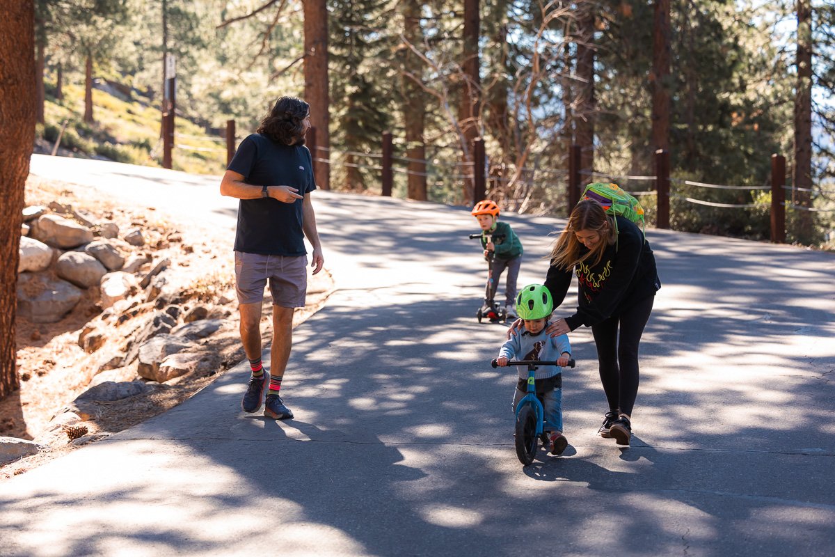 Mom steadying toddler on balance bike while dad walks alongside, Lake Tahoe neighborhood road, documentary family photography