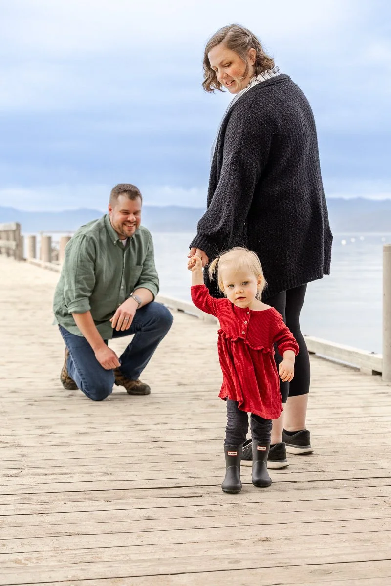 Toddler leading mom by the hand along the dock at Camp Richardson while dad watches during a documentary family photography session at Lake Tahoe