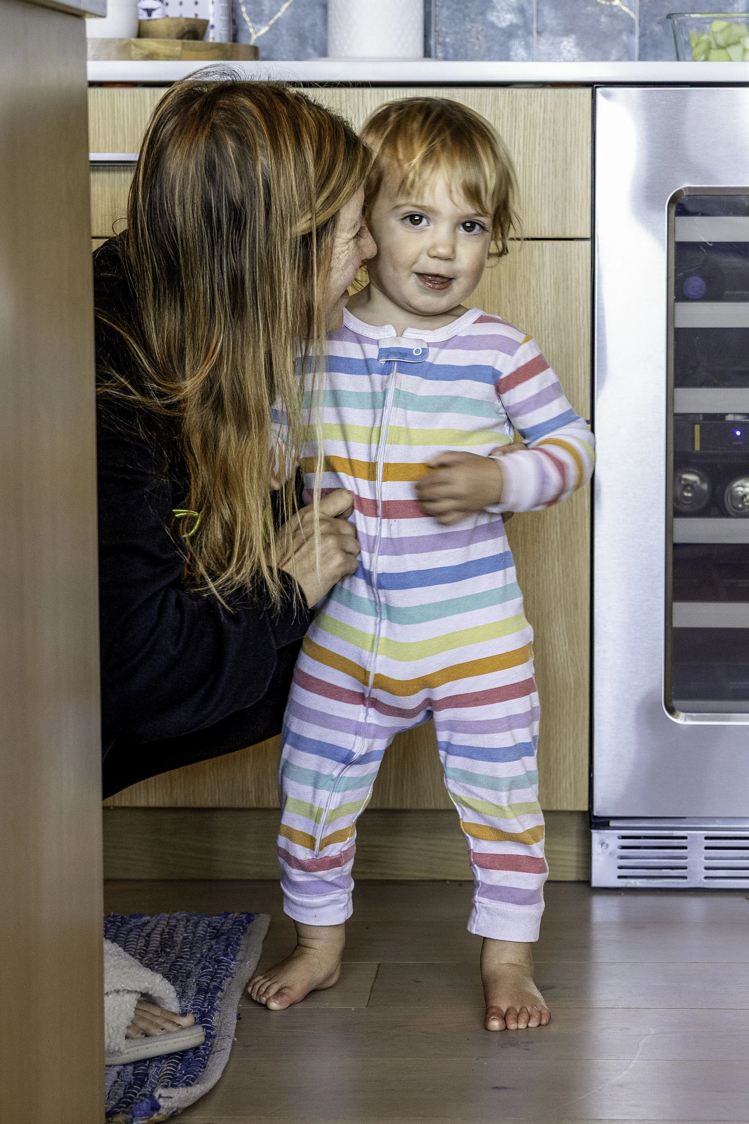 Mom kneeling on kitchen floor whispering to toddler in striped pajamas, Lake Tahoe cabin, documentary family photography