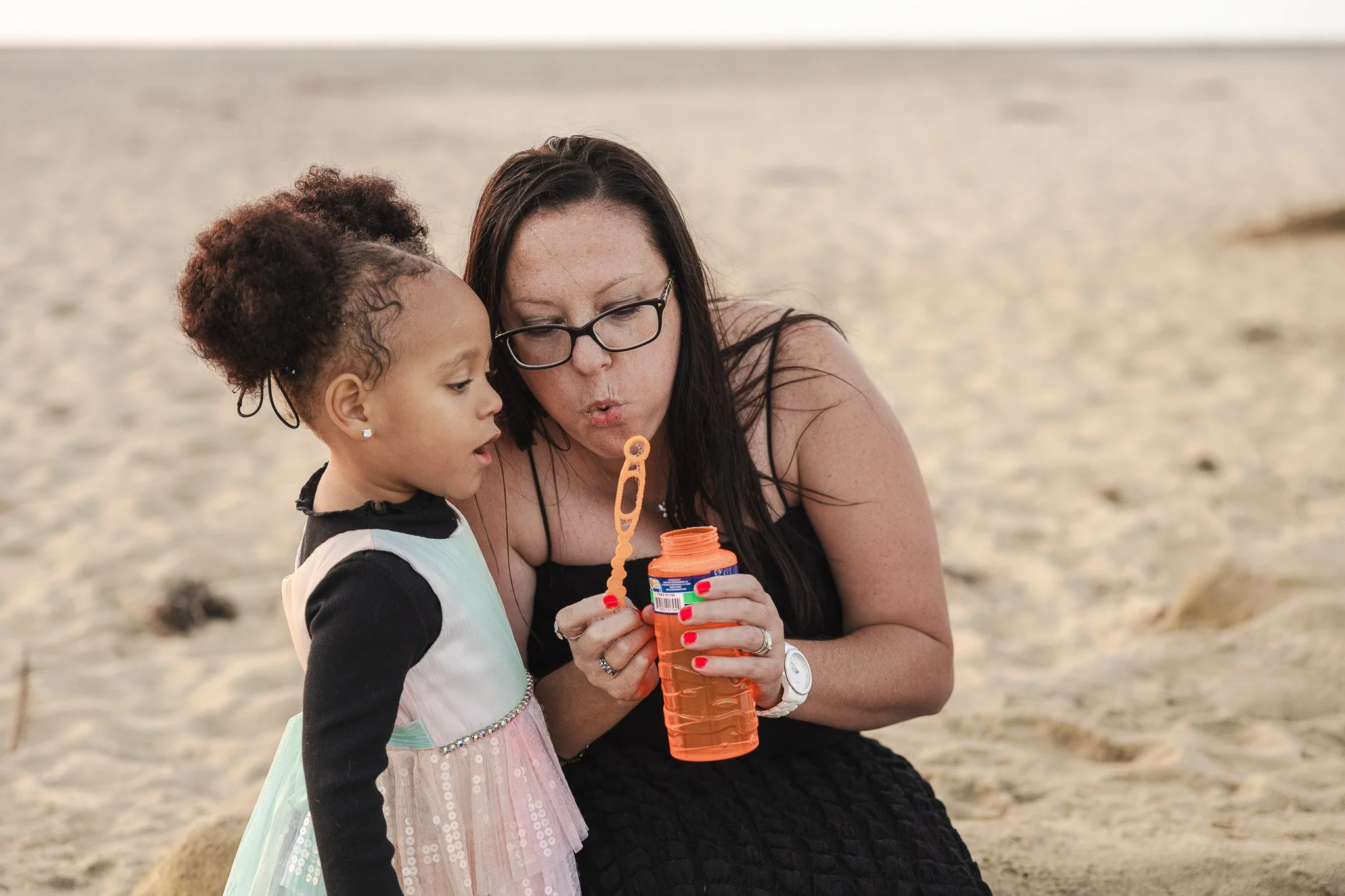 A mother crouches beside her young daughter on the beach, both focused on blowing bubbles together, a candid moment from a documentary family photography session by Ruwan & Diana Photography.