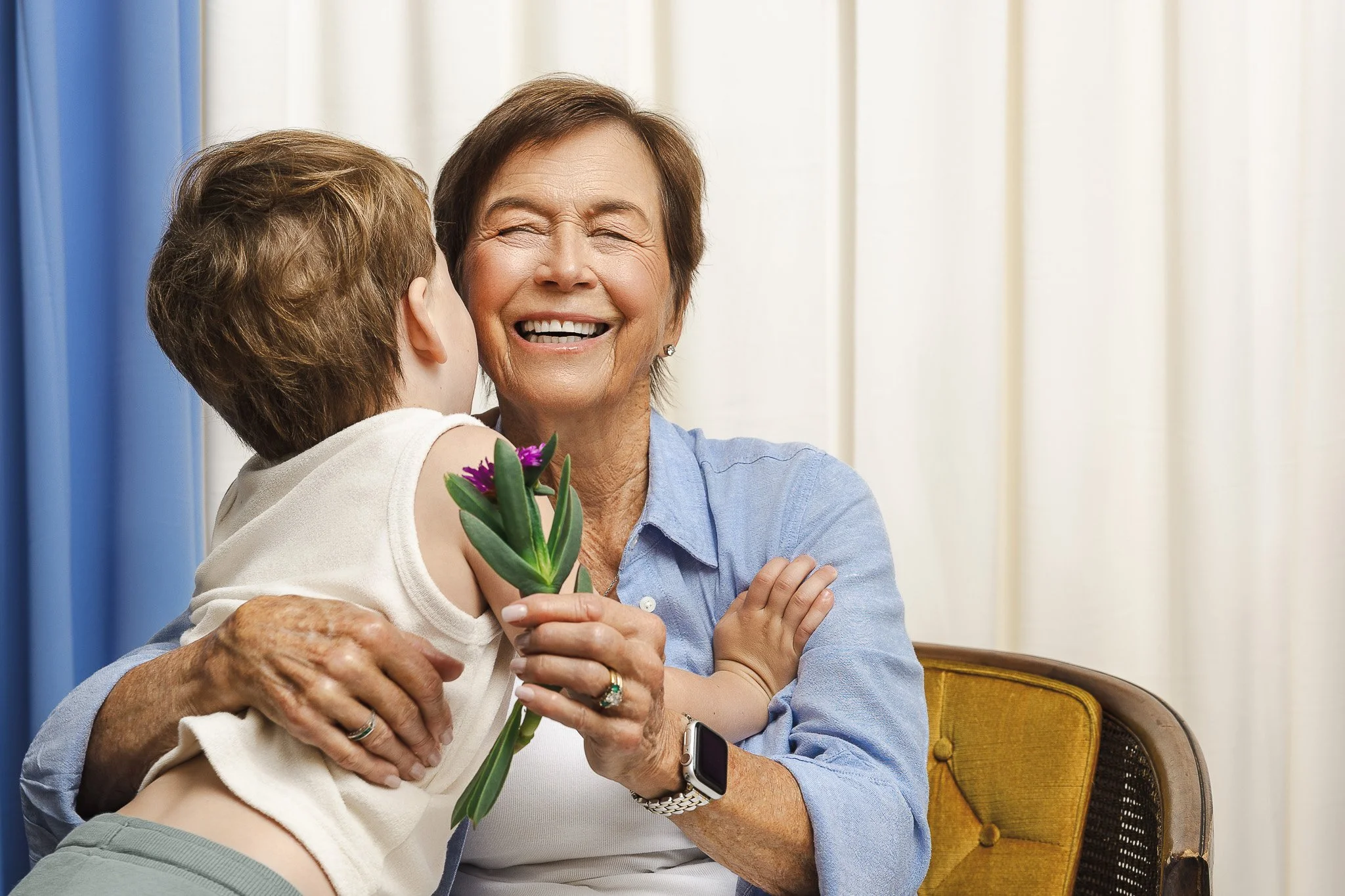 A young boy kisses his grandmother on the cheek while presenting her with a flower, her arms wrapped around him in a spontaneous embrace, a multigenerational documentary family portrait by Ruwan & Diana Photography, Del Mar.