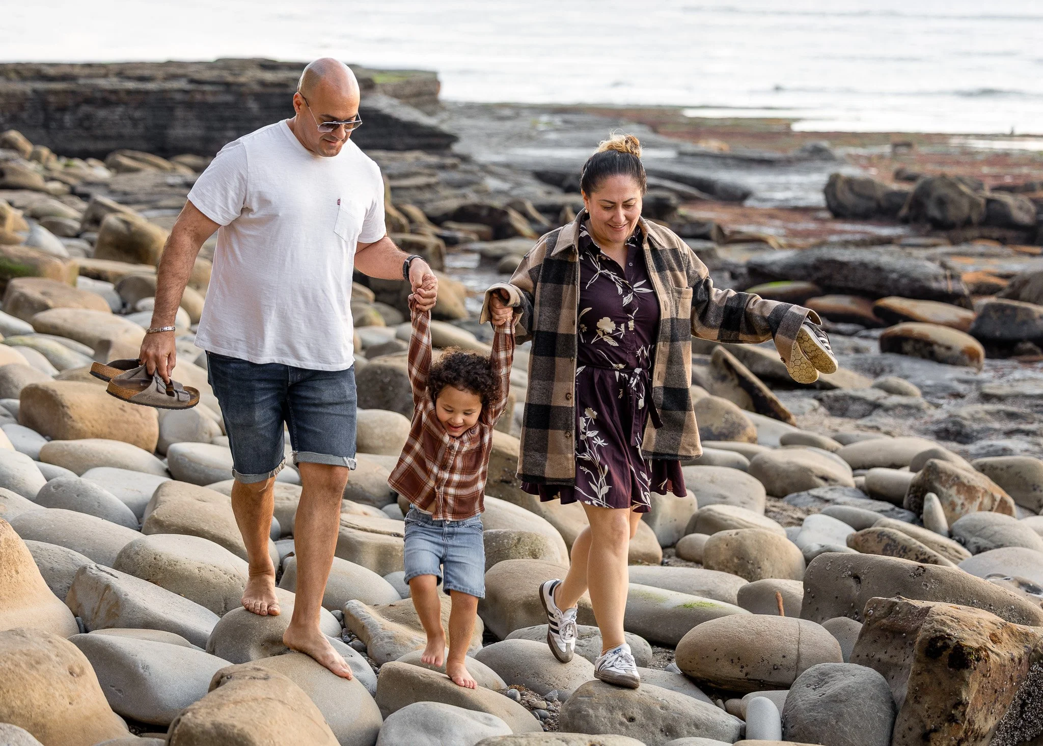A young child is swung between his parents as they navigate the rocky tidepools along the Southern California coast, a documentary family portrait by Ruwan & Diana Photography, Del Mar.