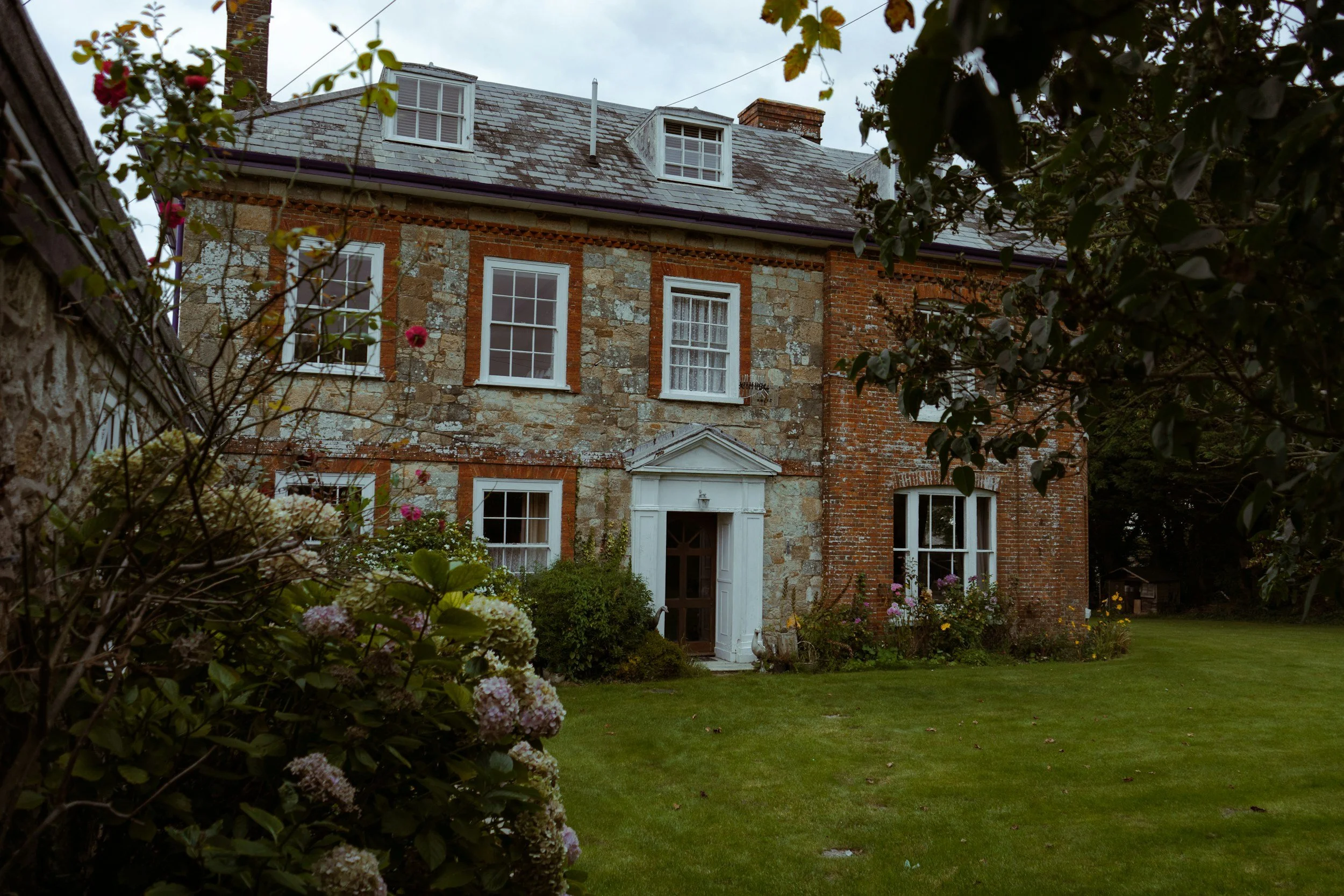 A historic brick and stone house with white-framed windows and a white door, surrounded by lush garden and flowering plants, under a cloudy sky.
