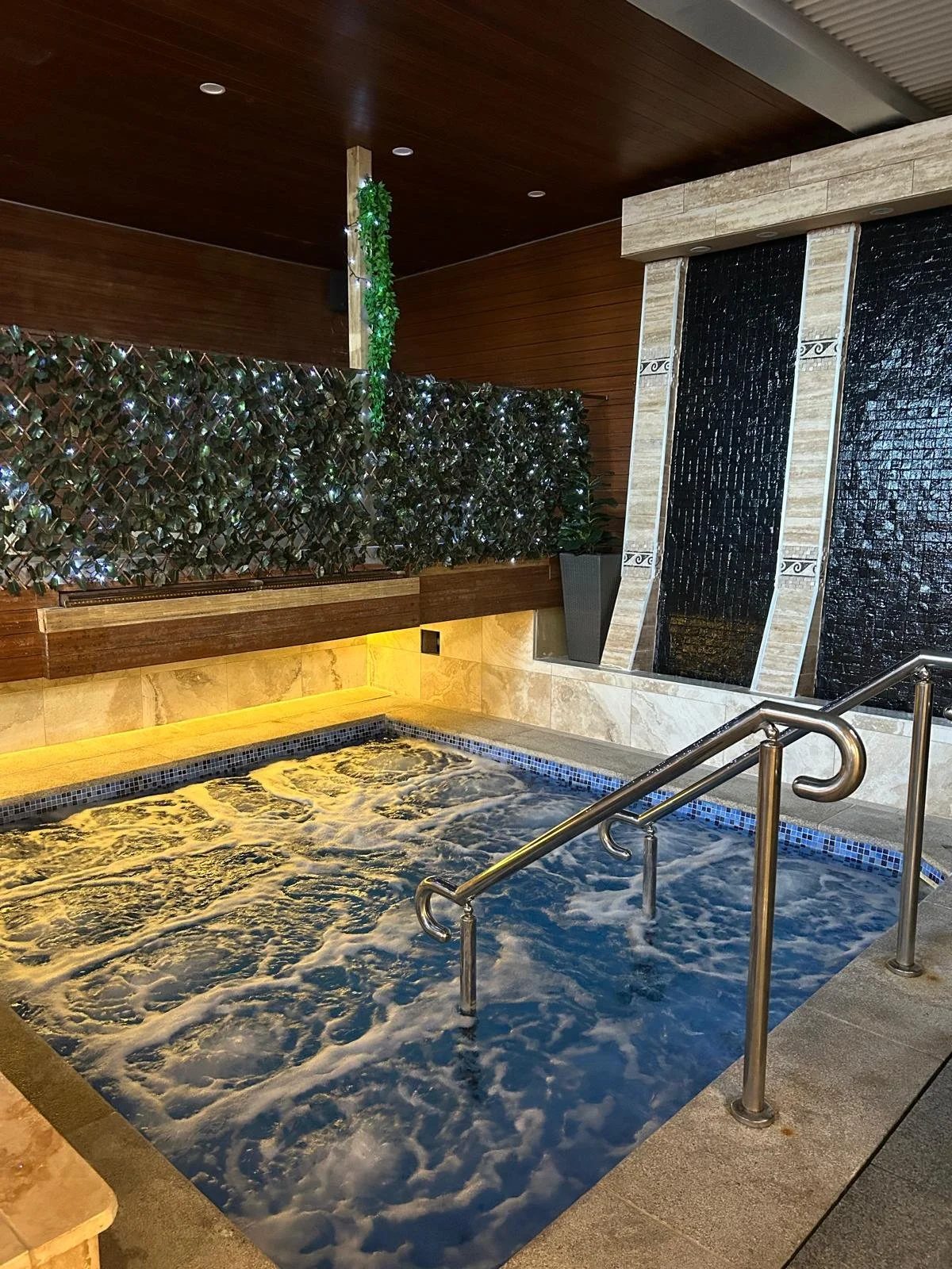 Indoor hot tub with bubbling water, stainless steel handrails, a wooden wall with decorative green plants and white string lights, and a waterfall feature with black water flowing down tiled panels.