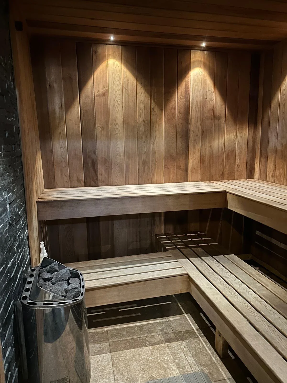 Wood-paneled sauna room with built-in benches, a heater with stones, and dim ceiling lighting.