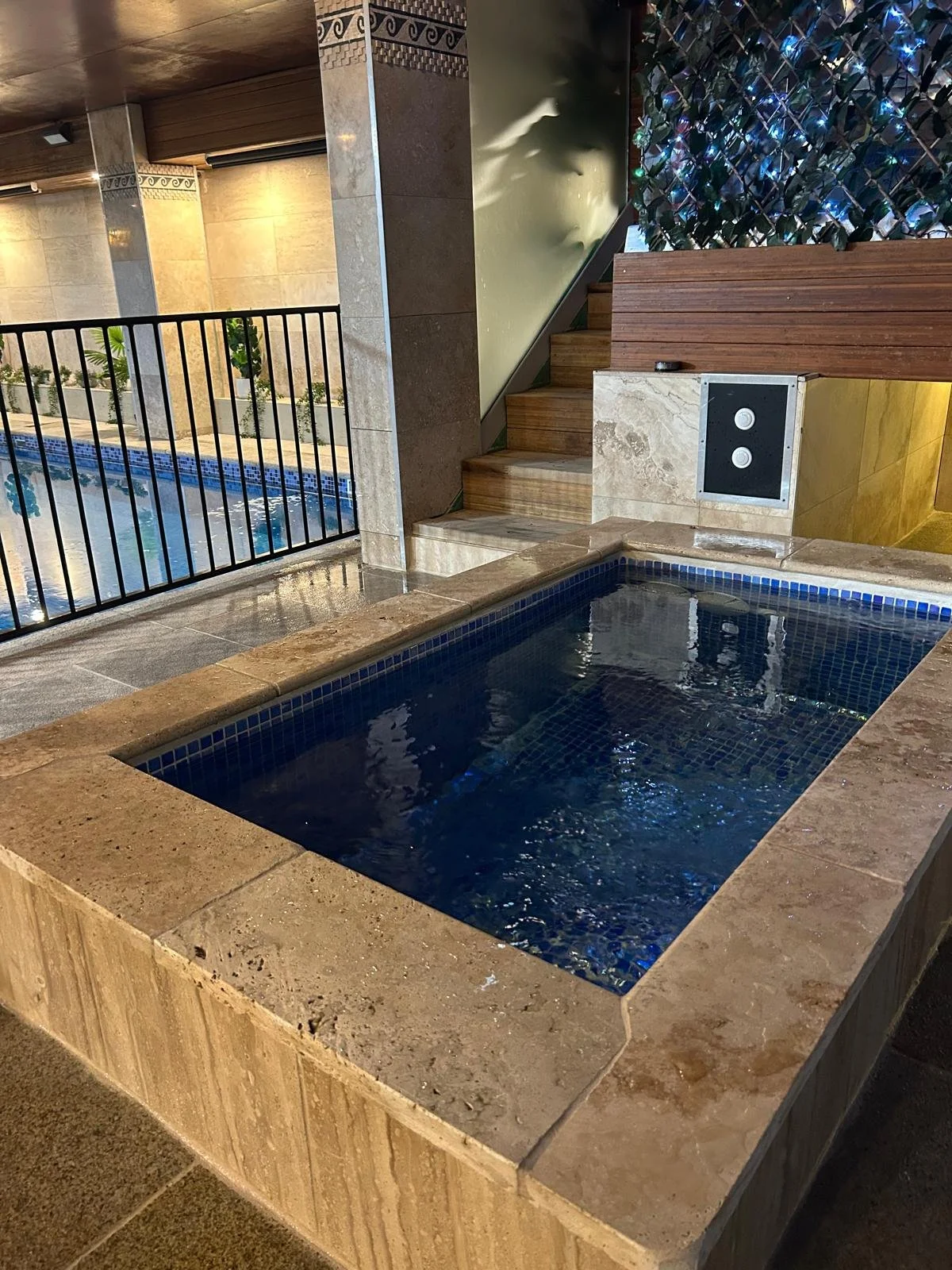 Indoor hot tub with dark blue water, surrounded by beige tile, near a staircase and a black railing, with plants and pool in the background.