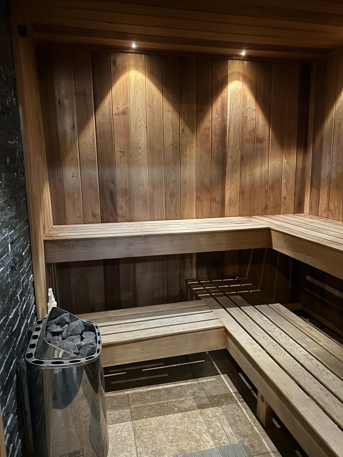 Wood-paneled sauna room with benches and a heater filled with rocks, illuminated by recessed lighting.